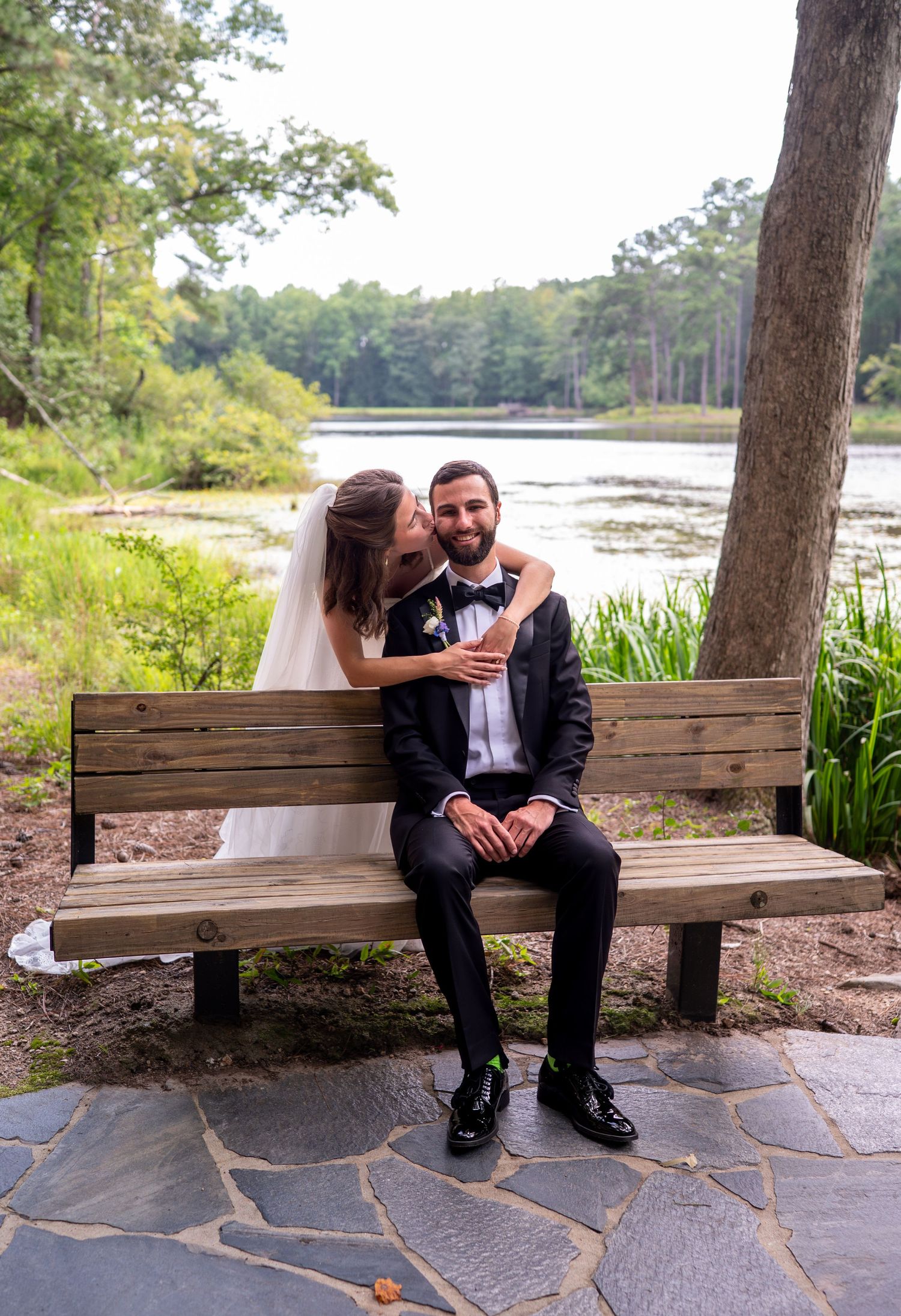 A couple shares a romantic moment on a wooden bench by a serene lake surrounded by trees on their wedding day.