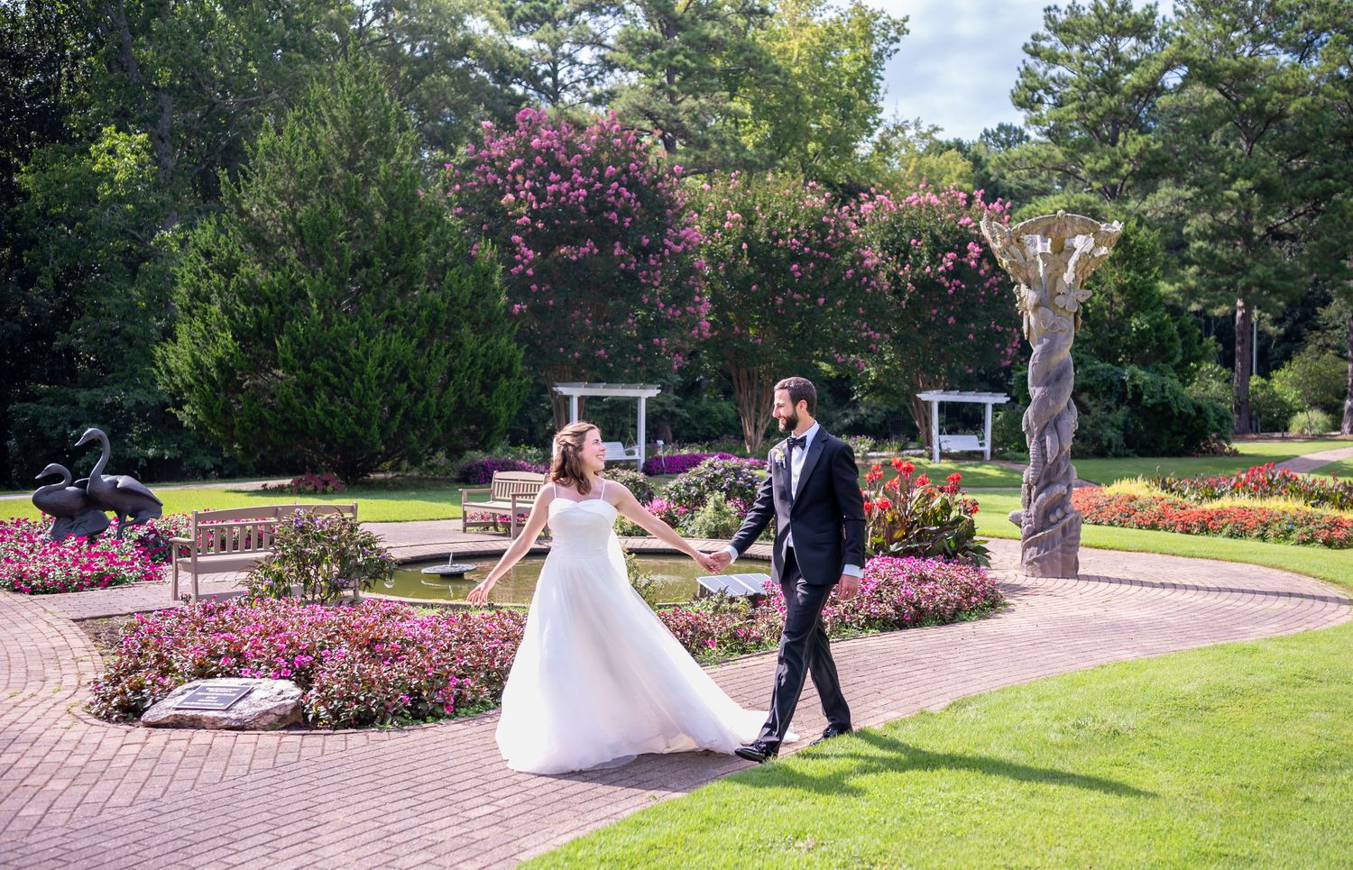 A romantic wedding photoshoot takes place in a lush garden setting with blooming pink flowers and manicured lawns.