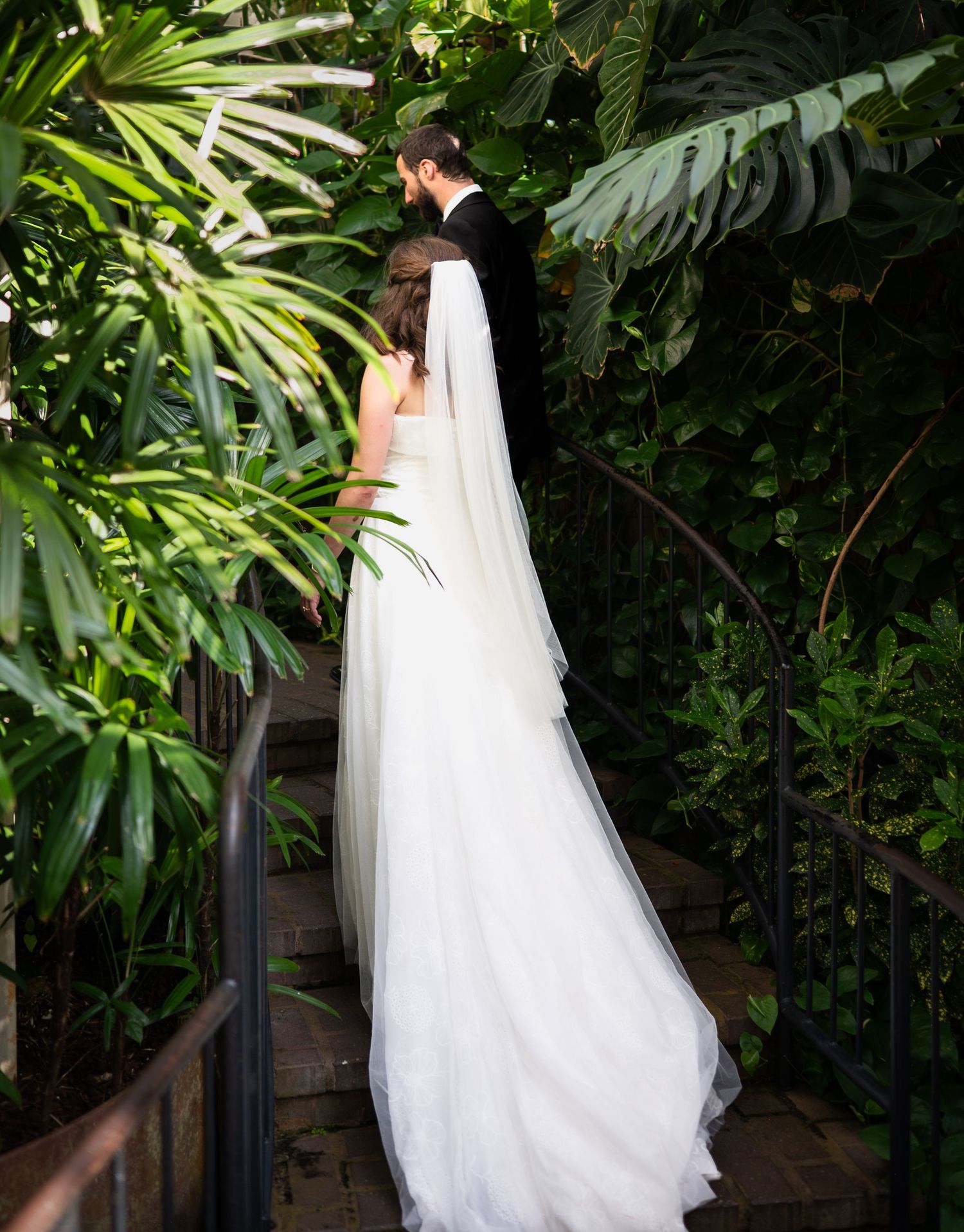 A flowing white wedding dress with a long train hangs elegantly against a backdrop of lush tropical greenery.