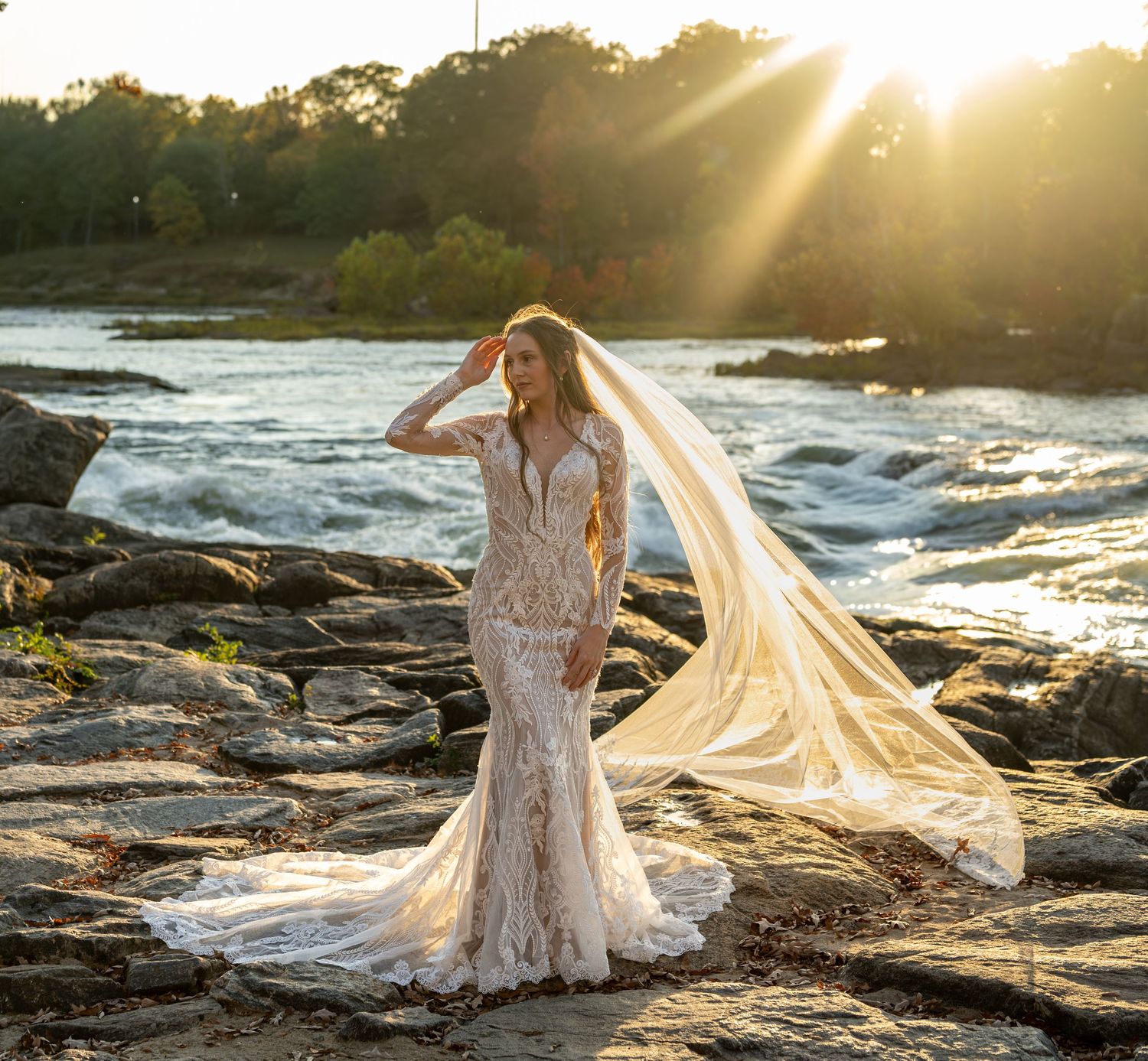 Dramatic sunset bridal portrait with flowing veil on rocky riverbank.
