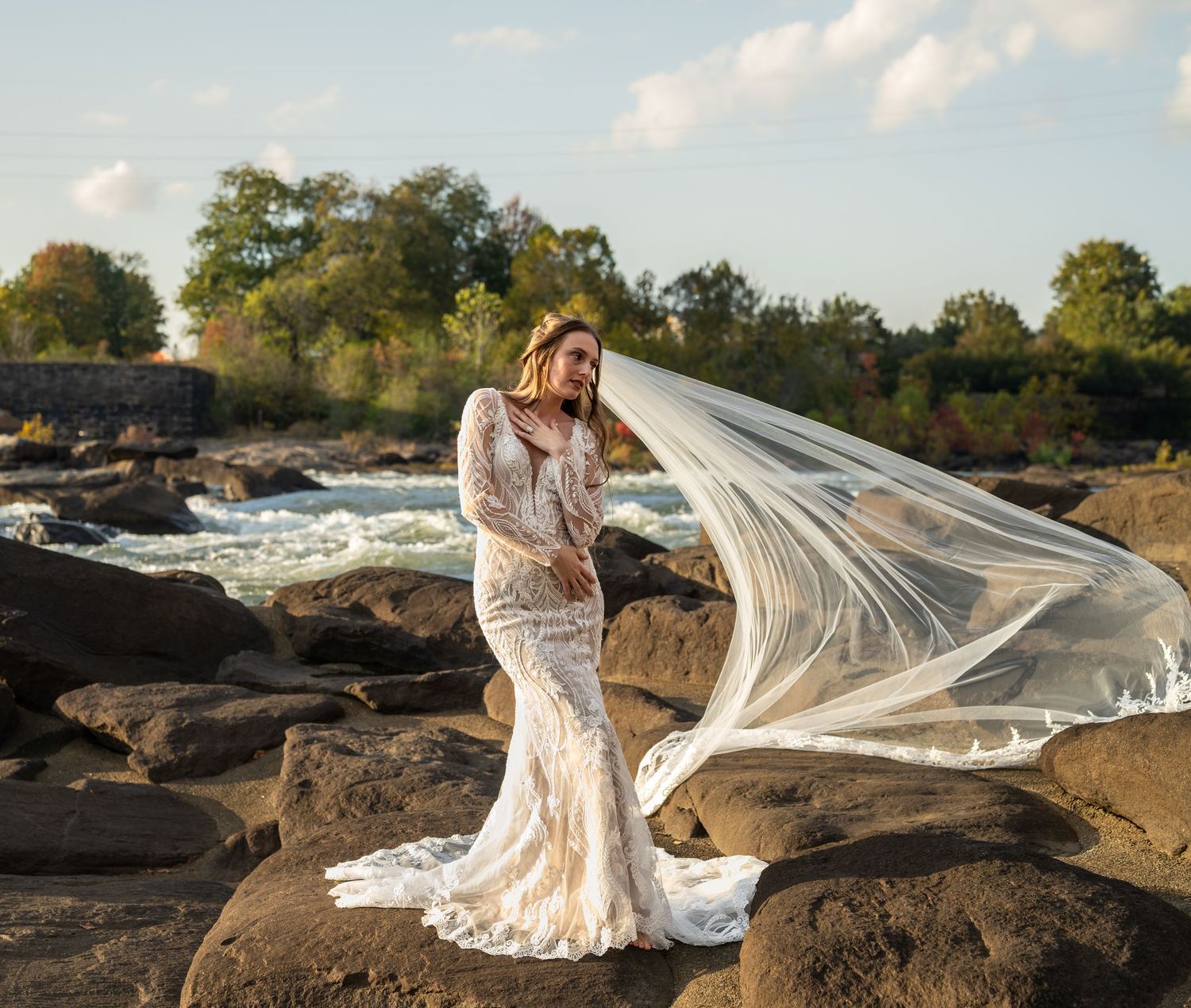 A flowing white wedding dress billows dramatically against a rocky coastal landscape at sunset.