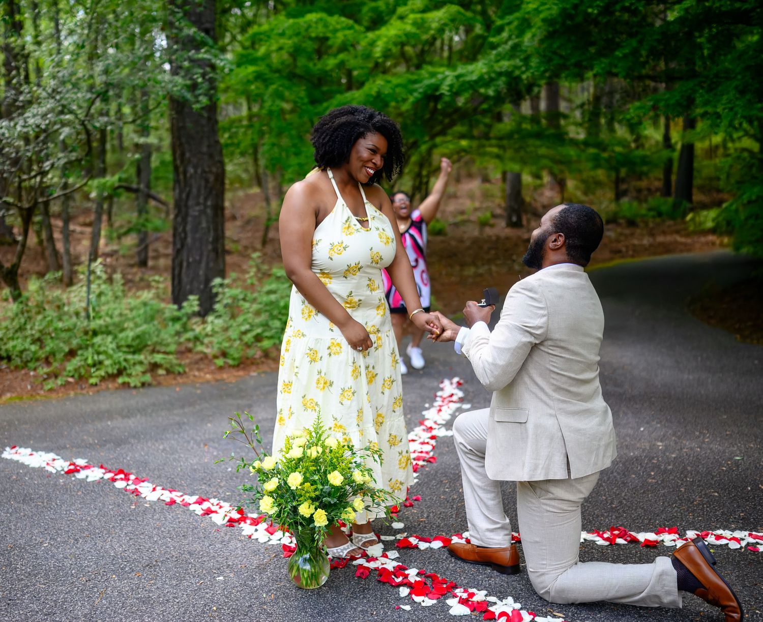 Man in white suit proposing on one knee to woman in yellow floral dress with excited guest celebrating behind.