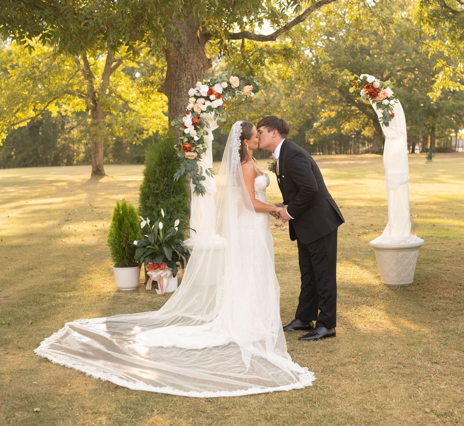 Romantic outdoor wedding ceremony setup with white floral arches and greenery in a sunlit garden setting.