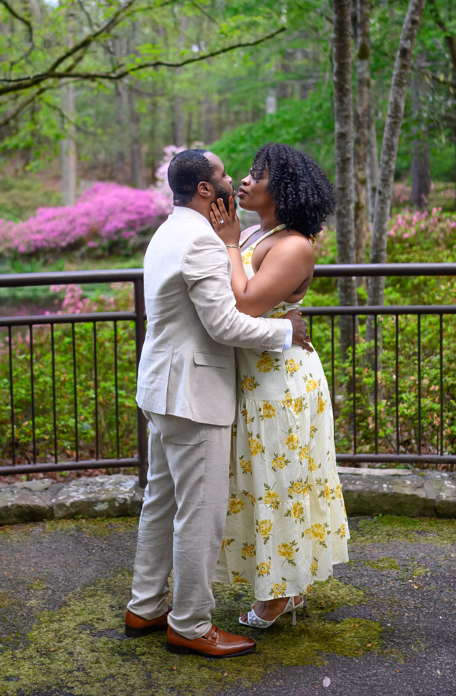 Romantic couple shares a kiss on a moss-covered patio surrounded by blooming azaleas in a garden.