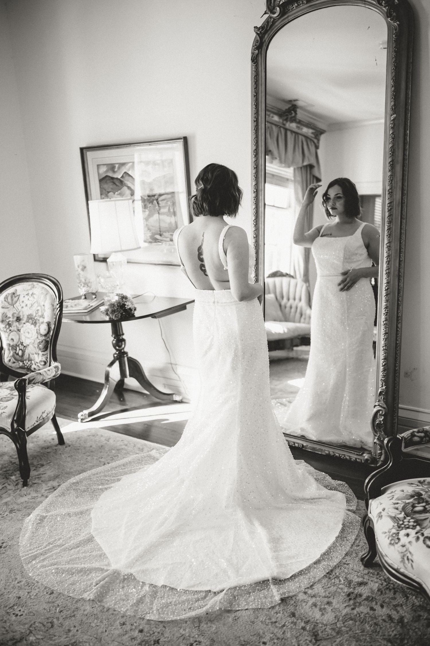 A bride in an elegant white lace wedding gown looks at her reflection in an ornate full-length mirror in a vintage room setting.
