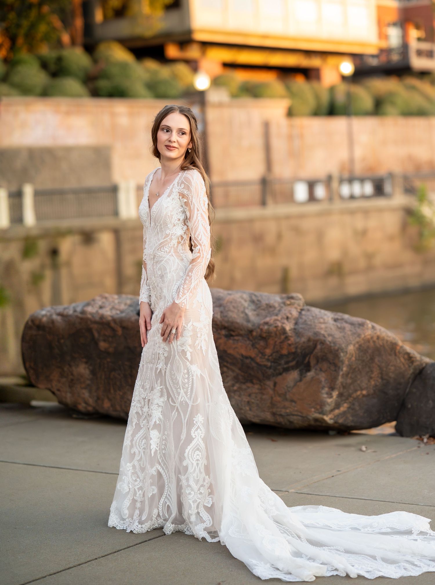 Elegant bride in fitted lace wedding dress poses by large rock during golden hour.