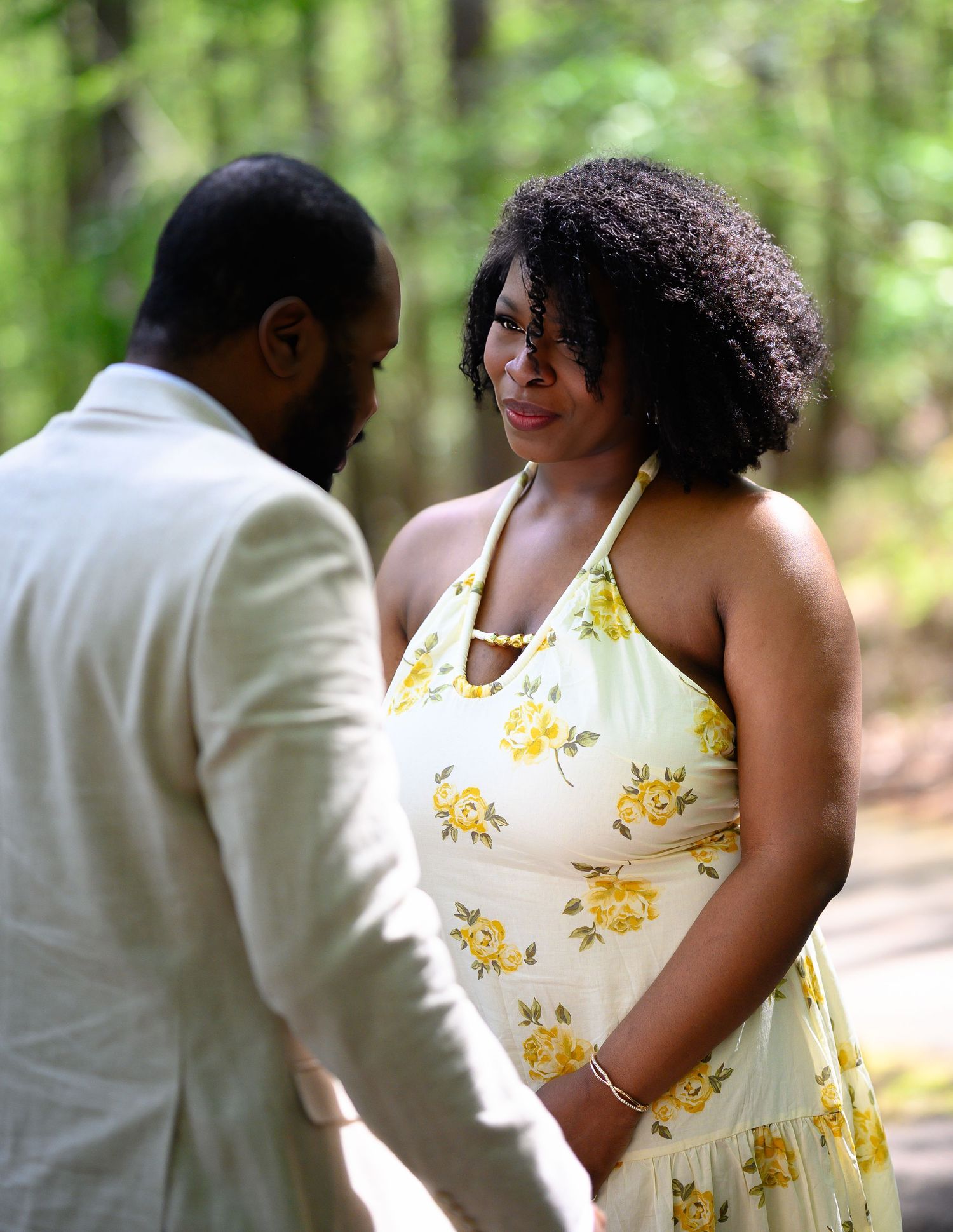 A couple shares a tender moment outdoors, woman in yellow floral dress, man in white suit, surrounded by trees.