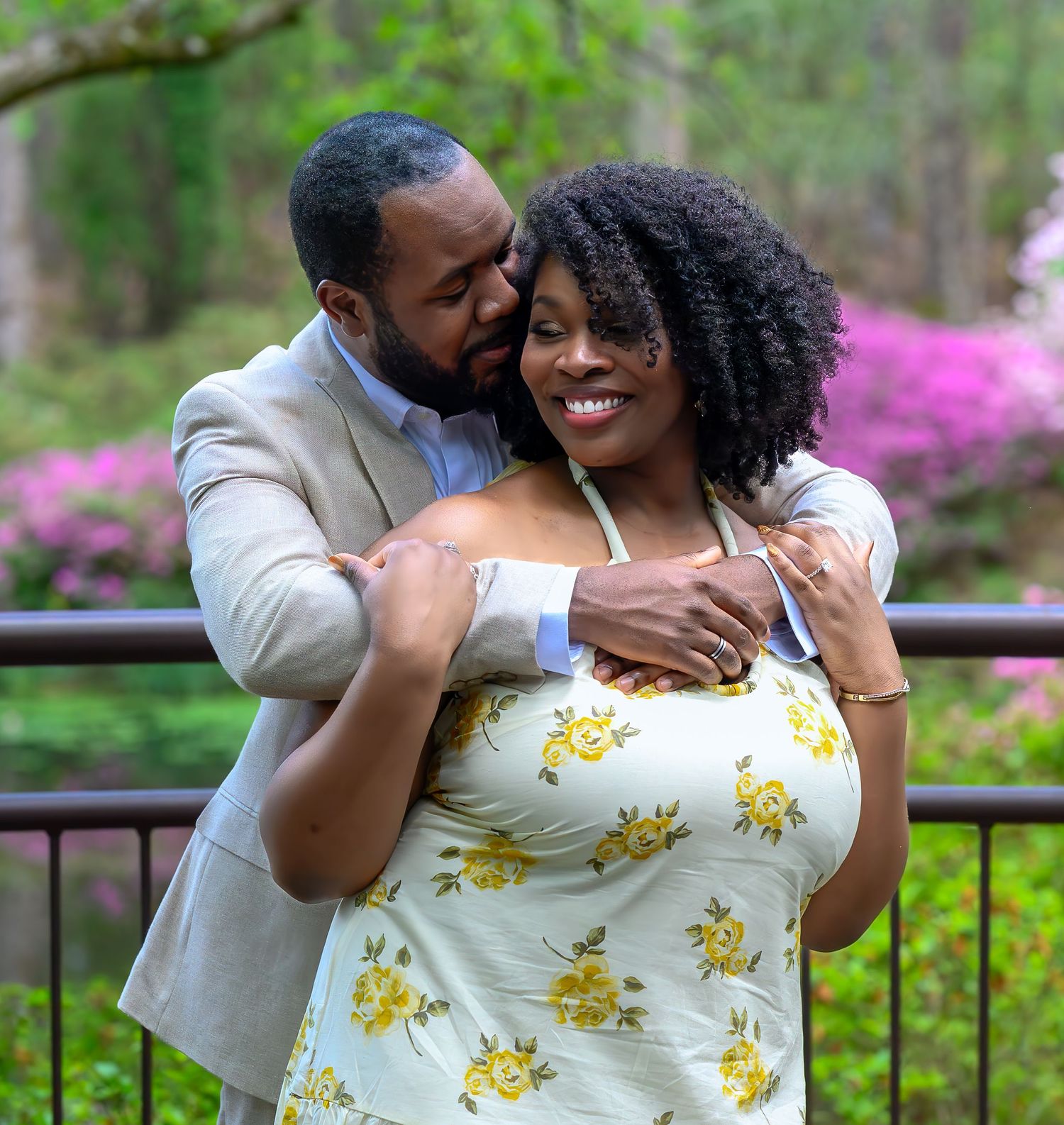 Happy couple embracing near a railing with blooming pink azaleas in a lush green garden backdrop.