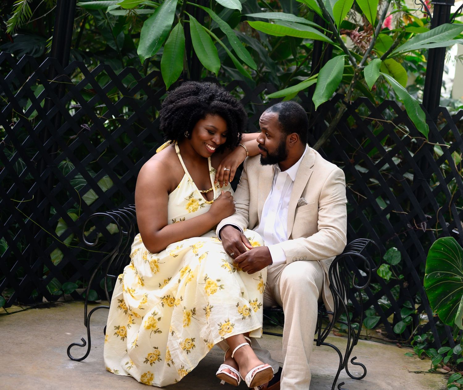 Engaged couple sits together on a bench surrounded by lush tropical plants in a garden setting.