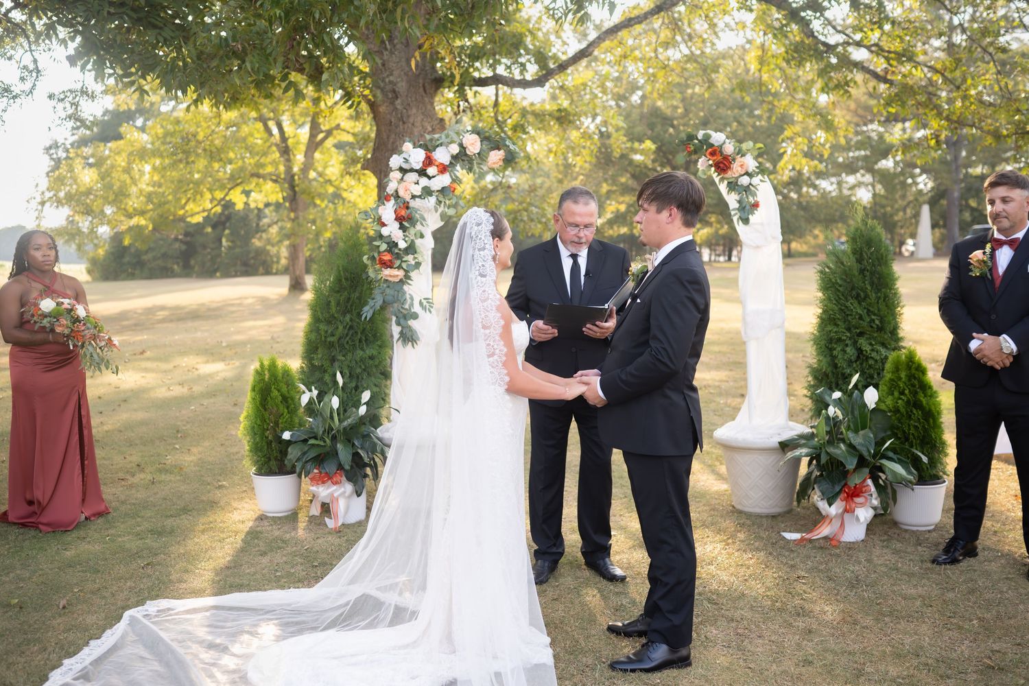 Outdoor wedding ceremony under trees with white decor and rust-colored bridesmaid dresses.