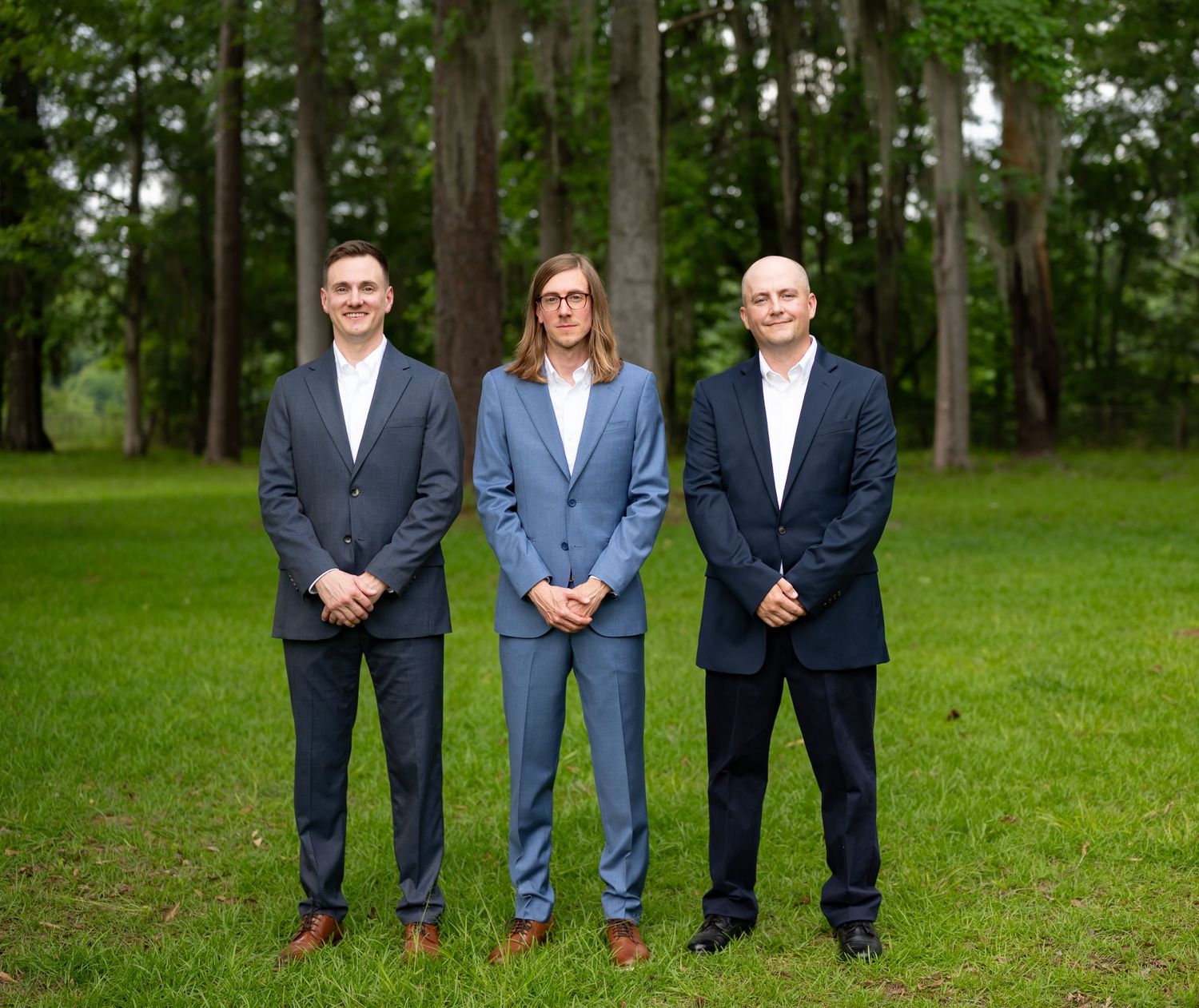 Row of groomsmen in navy suits pose formally on green lawn with trees in background.