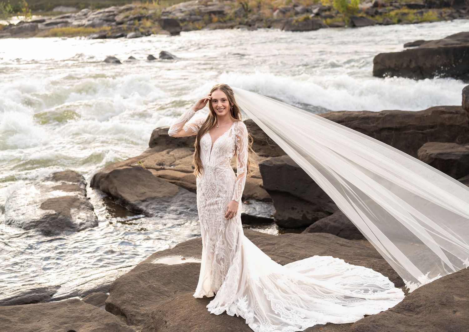 A flowing white wedding dress with lace details trails gracefully across dark coastal rocks near crashing ocean waves.