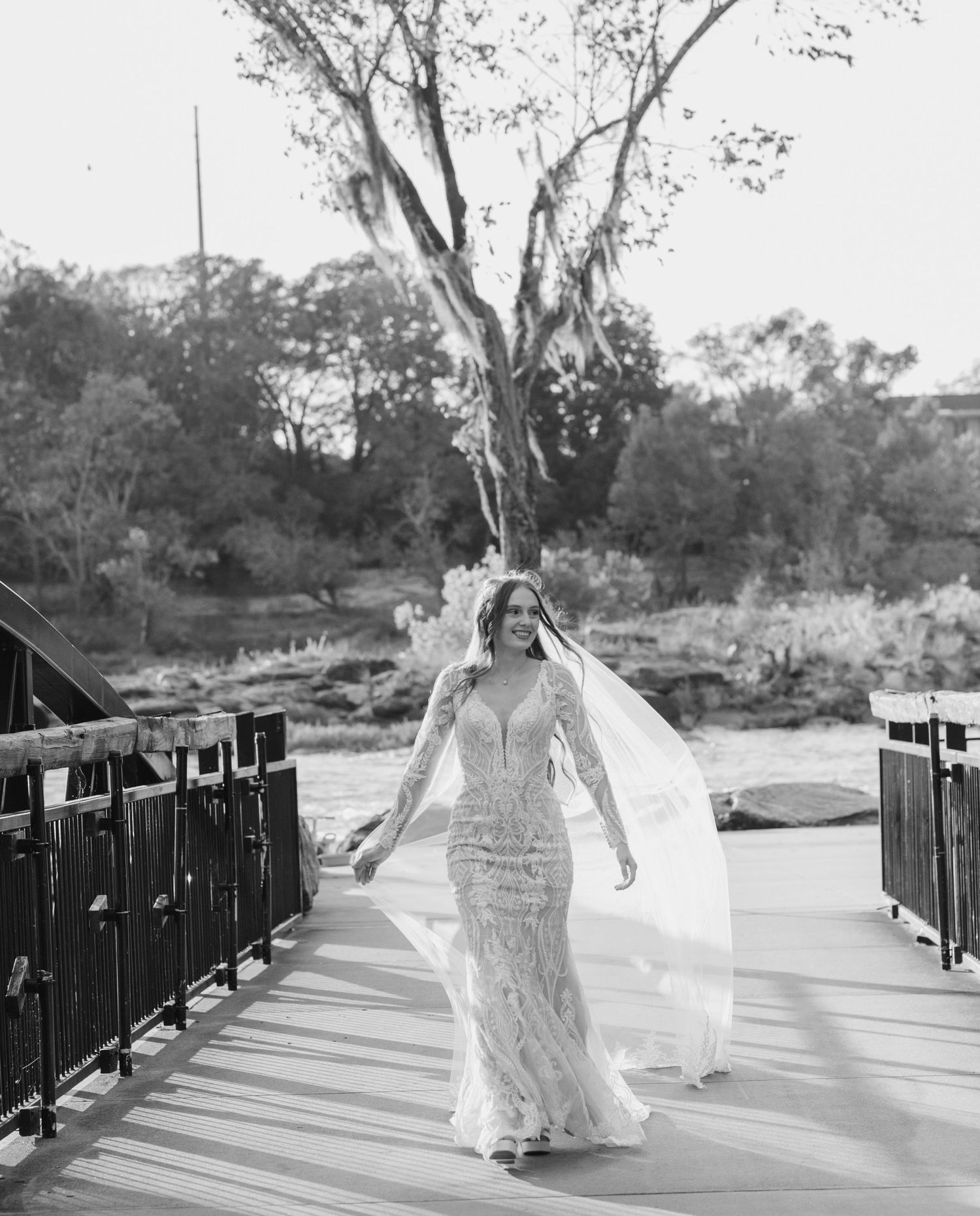 Black and white photo of a bride walking along a bridge railing in a flowing dress.