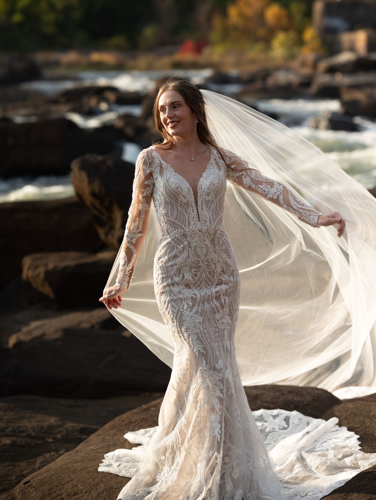 Elegant bride in lace wedding dress with flowing veil poses against rocky coastal backdrop at sunset.