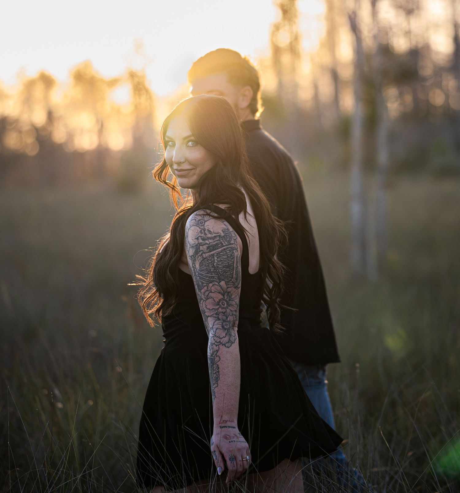 Person in black coat stands in golden sunlit field during sunset with backlit hair blowing in the wind.
