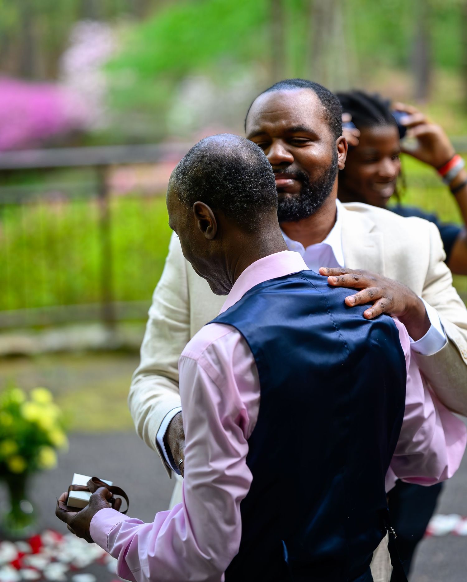 Two men embracing and celebrating outdoors near a garden with blooming azaleas in the background.