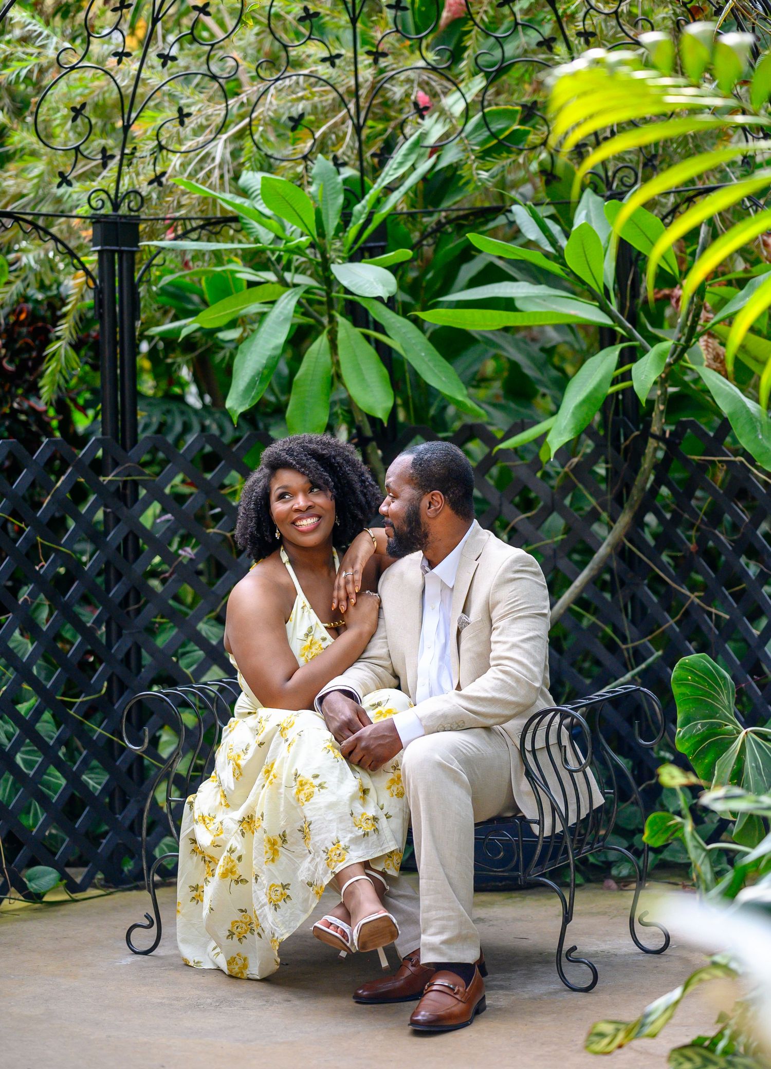 Couple sitting on ornate iron bench surrounded by tropical plants, sharing a loving glance and smiling.