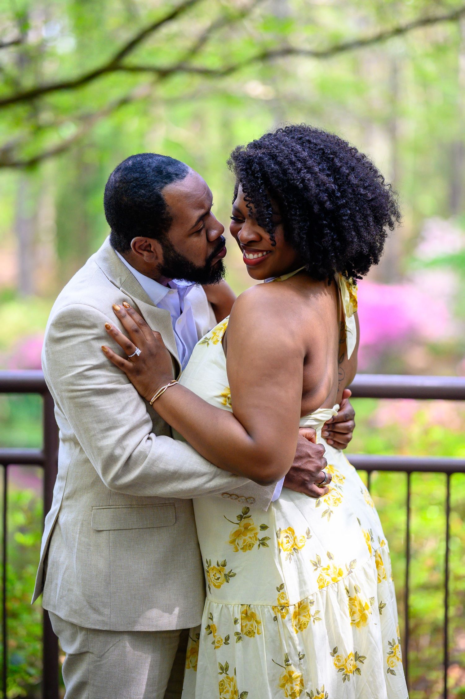Couple embracing and smiling intimately outdoors with pink flowering trees and wooden fence in background.