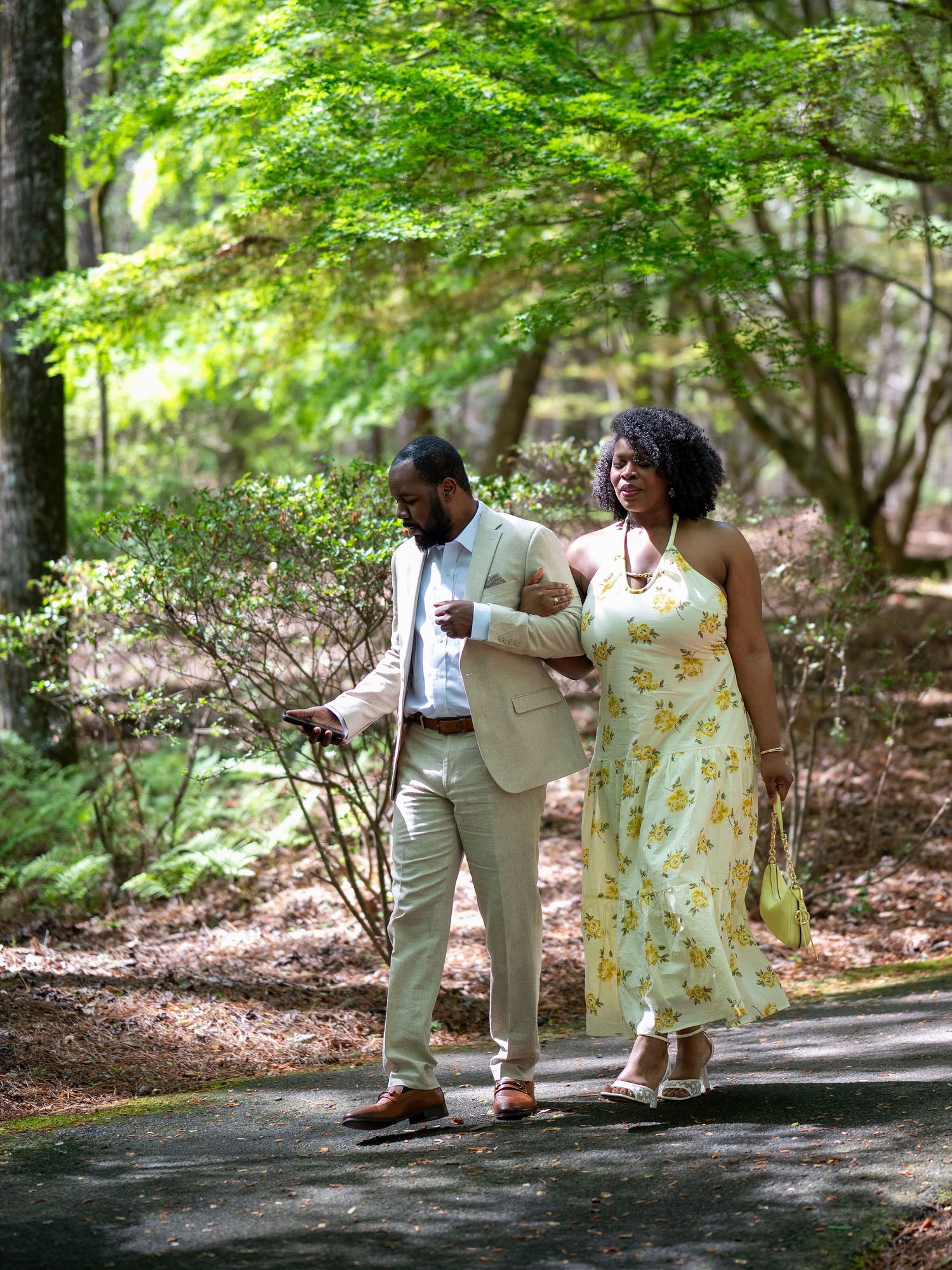 Couple walks hand-in-hand along a shaded forest path, man in tan suit, woman in yellow floral dress.