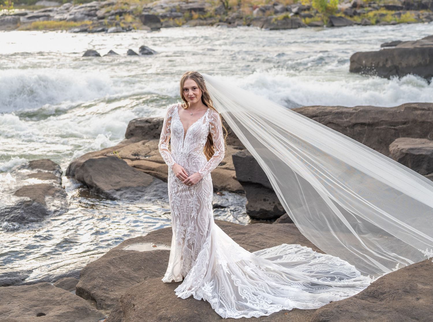 A flowing white lace wedding dress with long train photographed against dramatic coastal rocks and ocean waves.