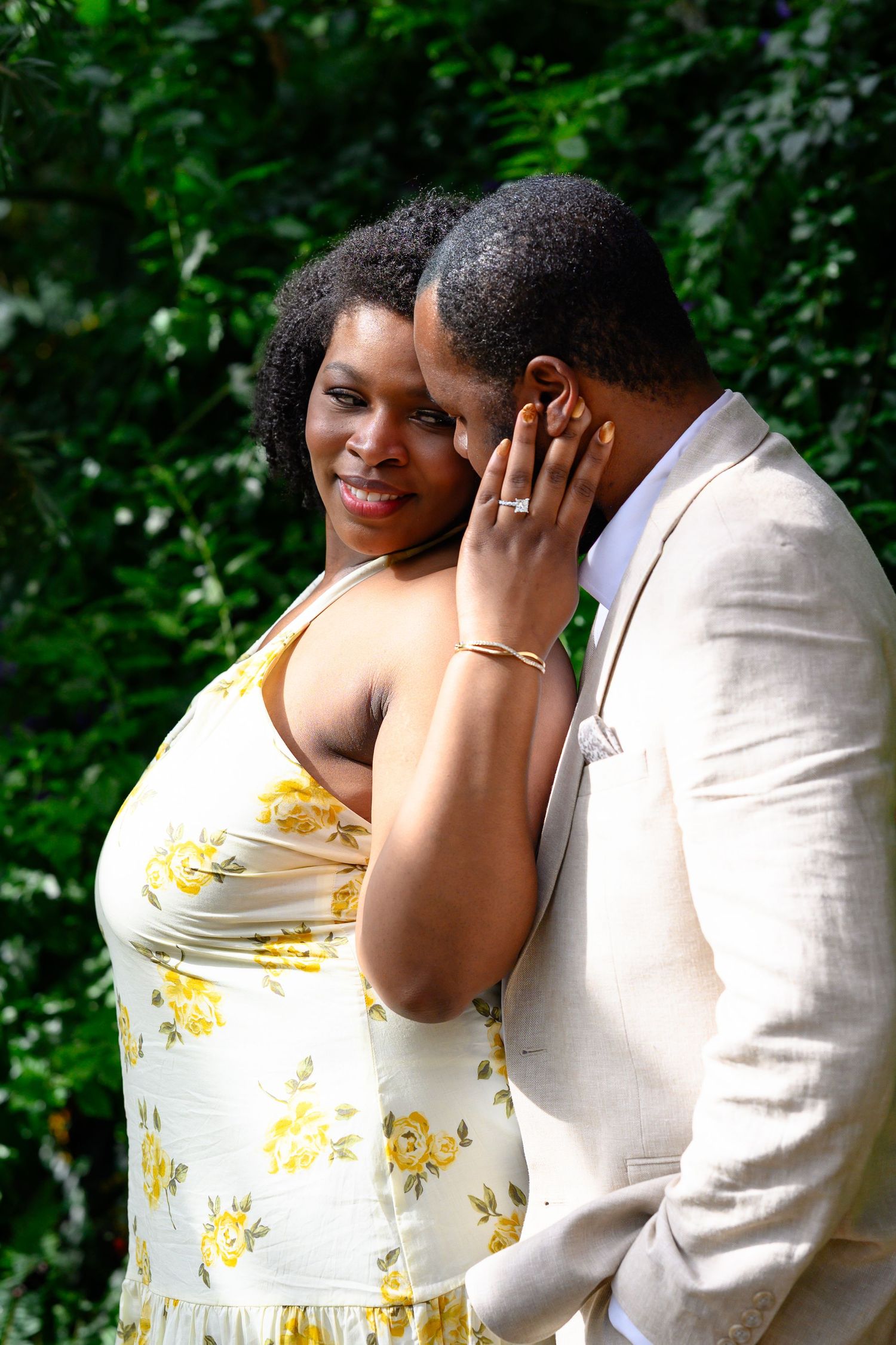 A woman smiles while gently touching a mans face outdoors with vibrant green foliage in the background.