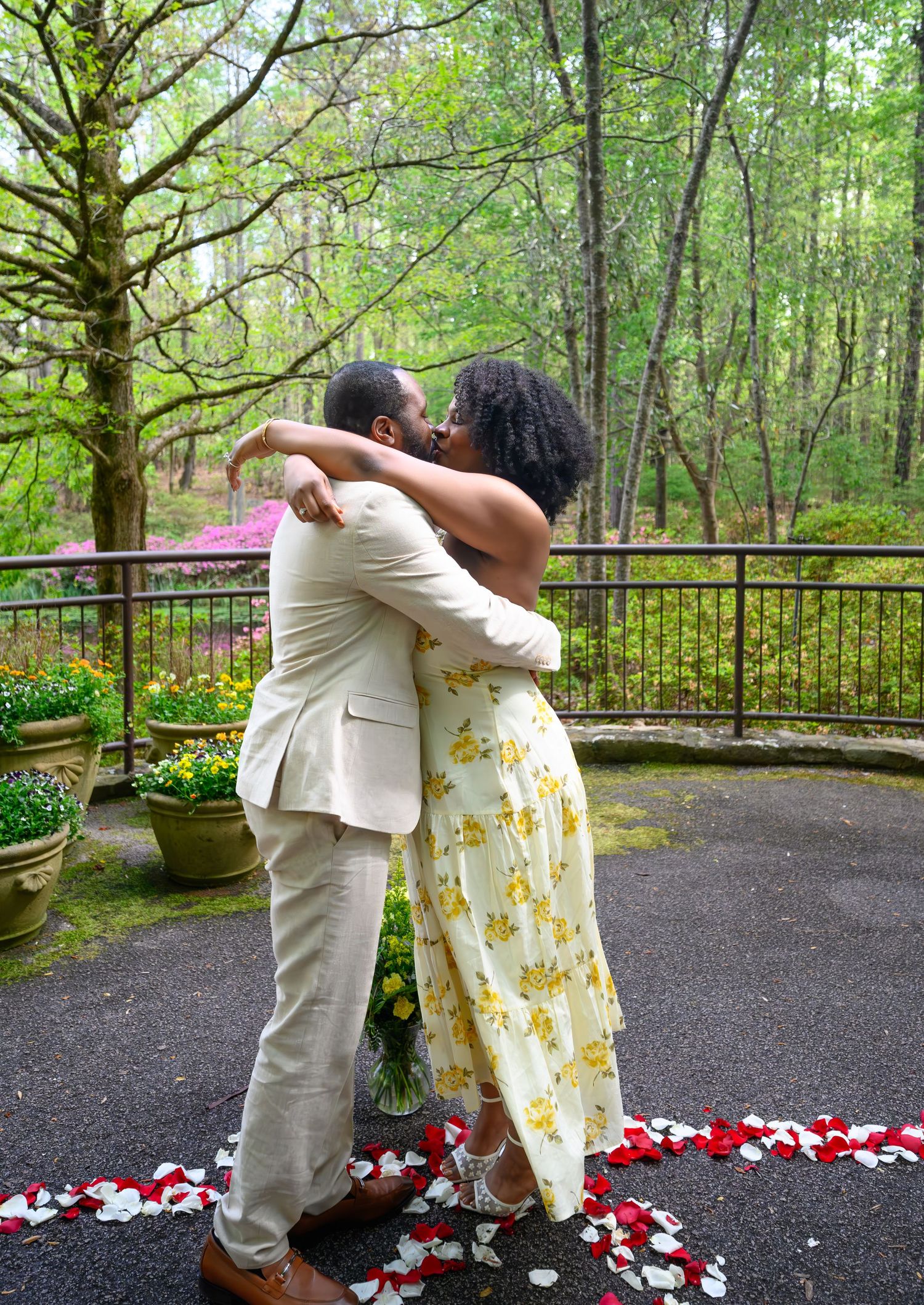 A joyful couple embraces and kisses outdoors surrounded by rose petals on the ground with a lush green forest backdrop.