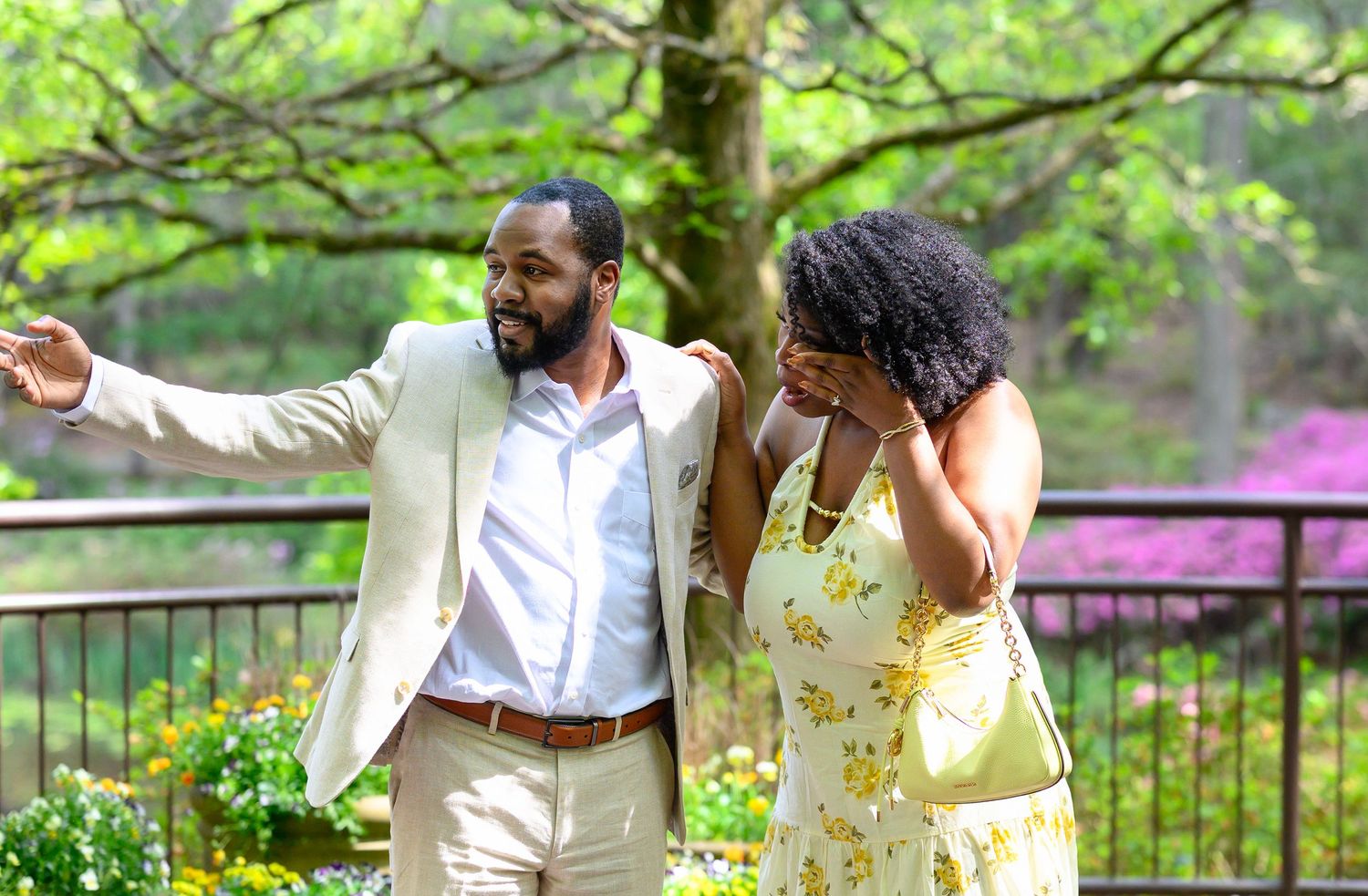 A man in a beige suit gestures while embracing a woman in a yellow floral dress outdoors.