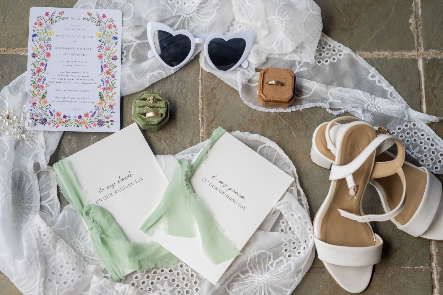 Delicate wedding details including floral invitations, silk ribbons, and elegant shoes arranged on white fabric background.