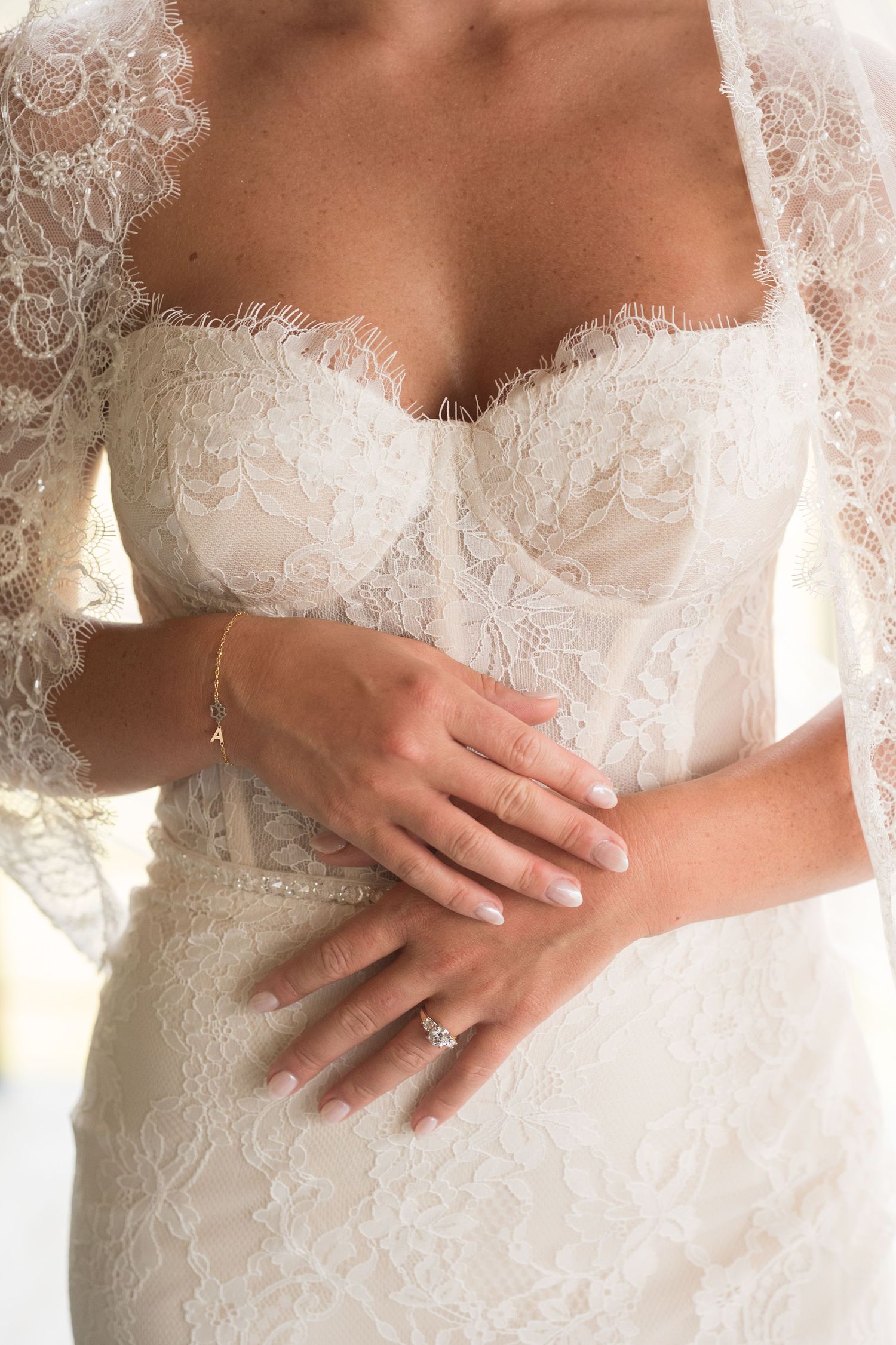 Close-up view of hands wearing an engagement ring against delicate white lace wedding dress details.