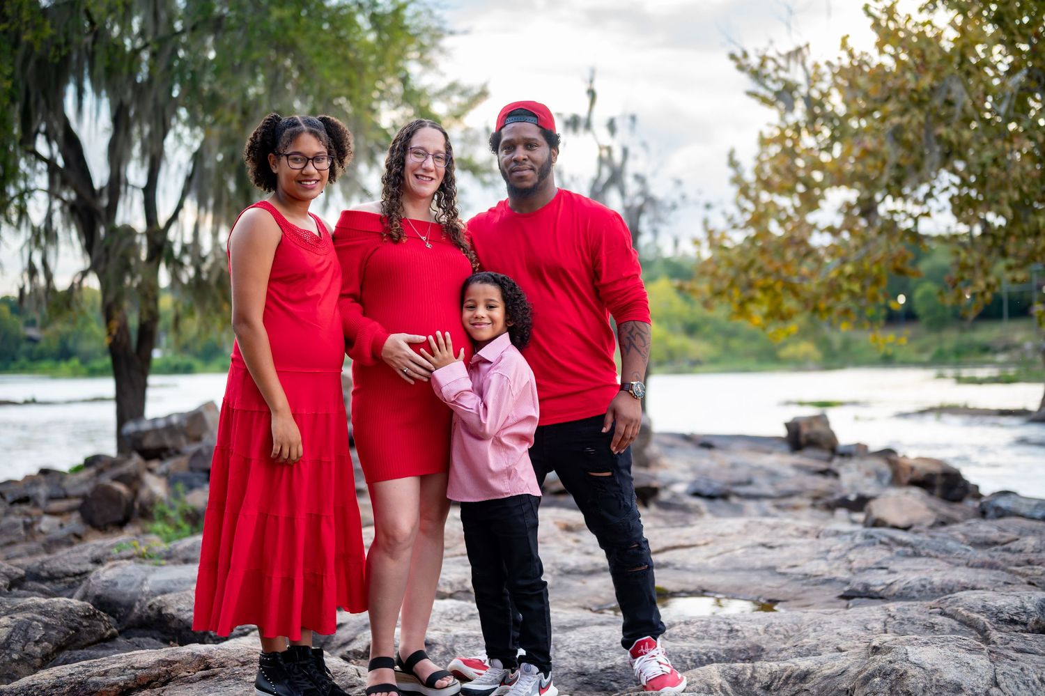 Family in coordinated red outfits smiles together on rocky waterfront during autumn photo session.
