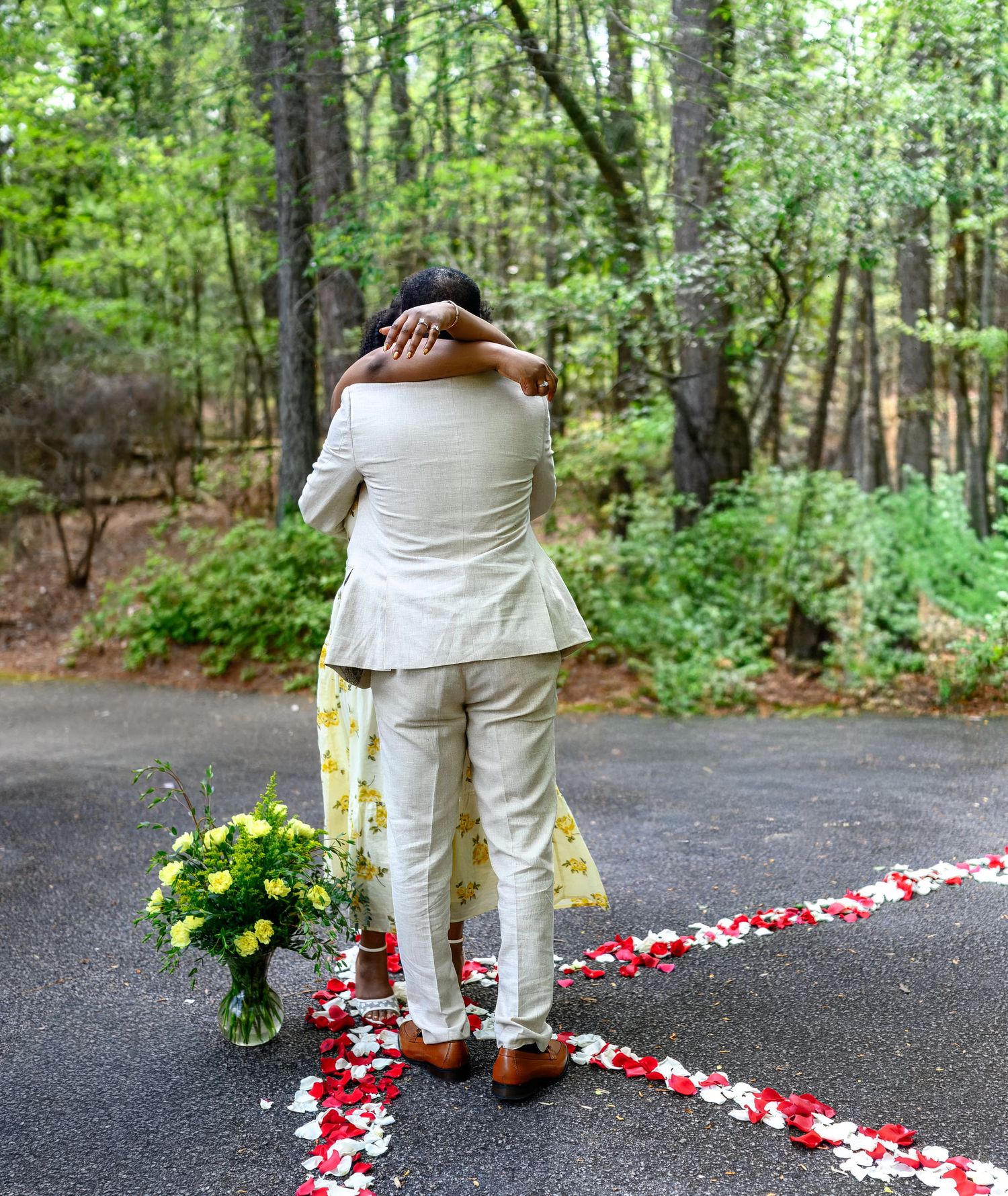 Newly engaged couple embraces on a petal-lined path surrounded by forest trees with a bouquet of yellow roses nearby.