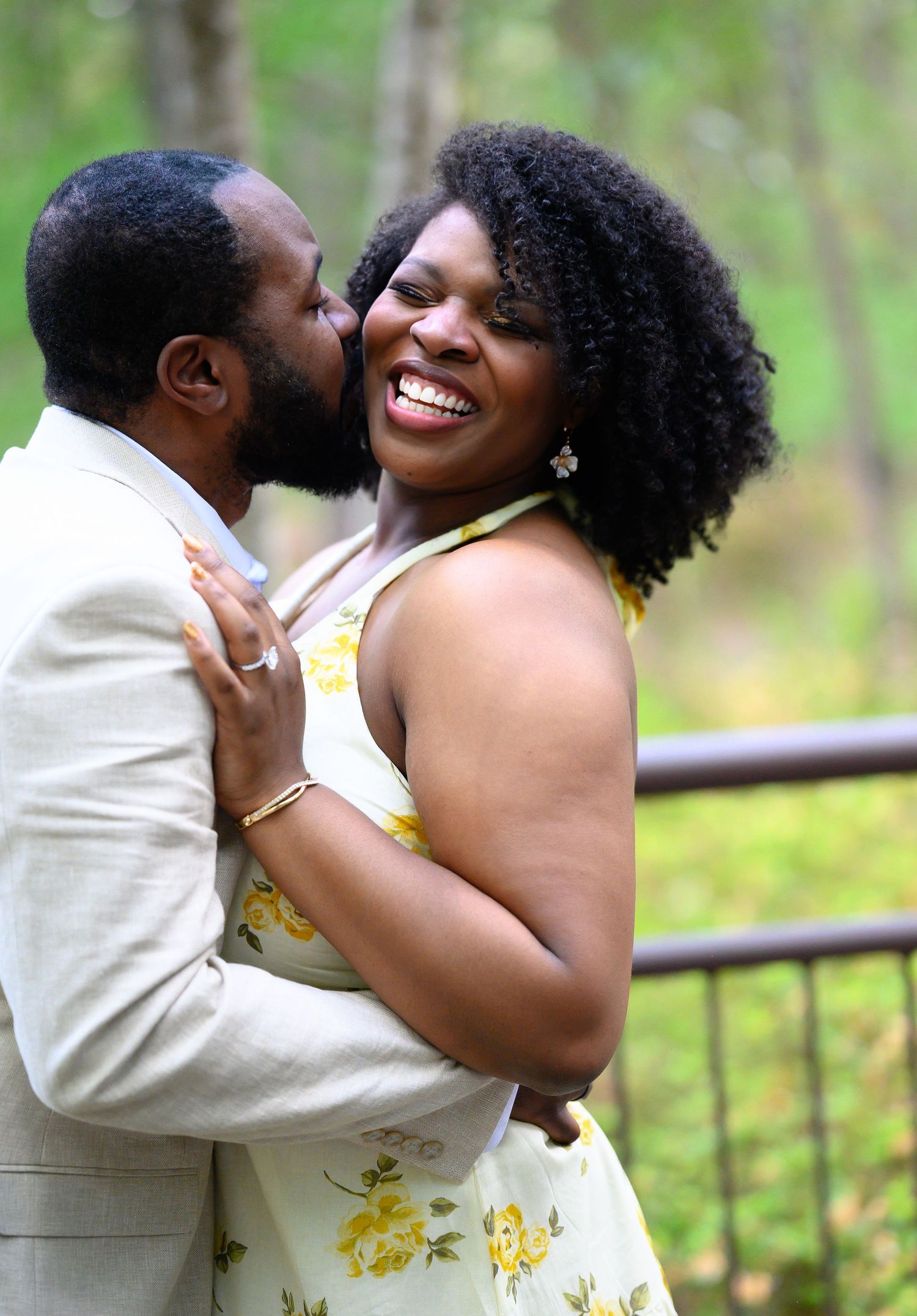 A joyful couple embraces outdoors, man in beige suit kisses womans cheek as she laughs in yellow floral dress.