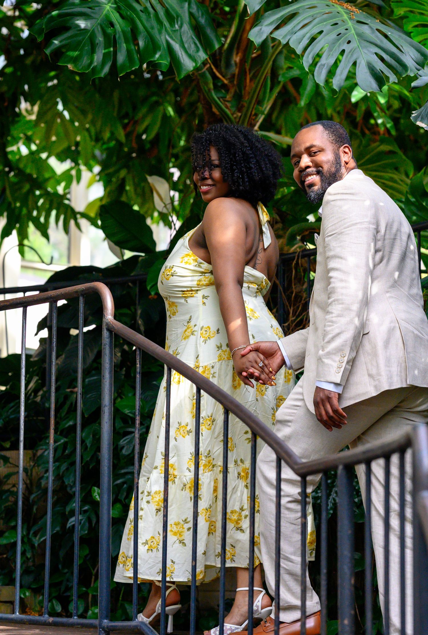 Happy couple holds hands and smiles while walking up stairs surrounded by tropical green foliage.