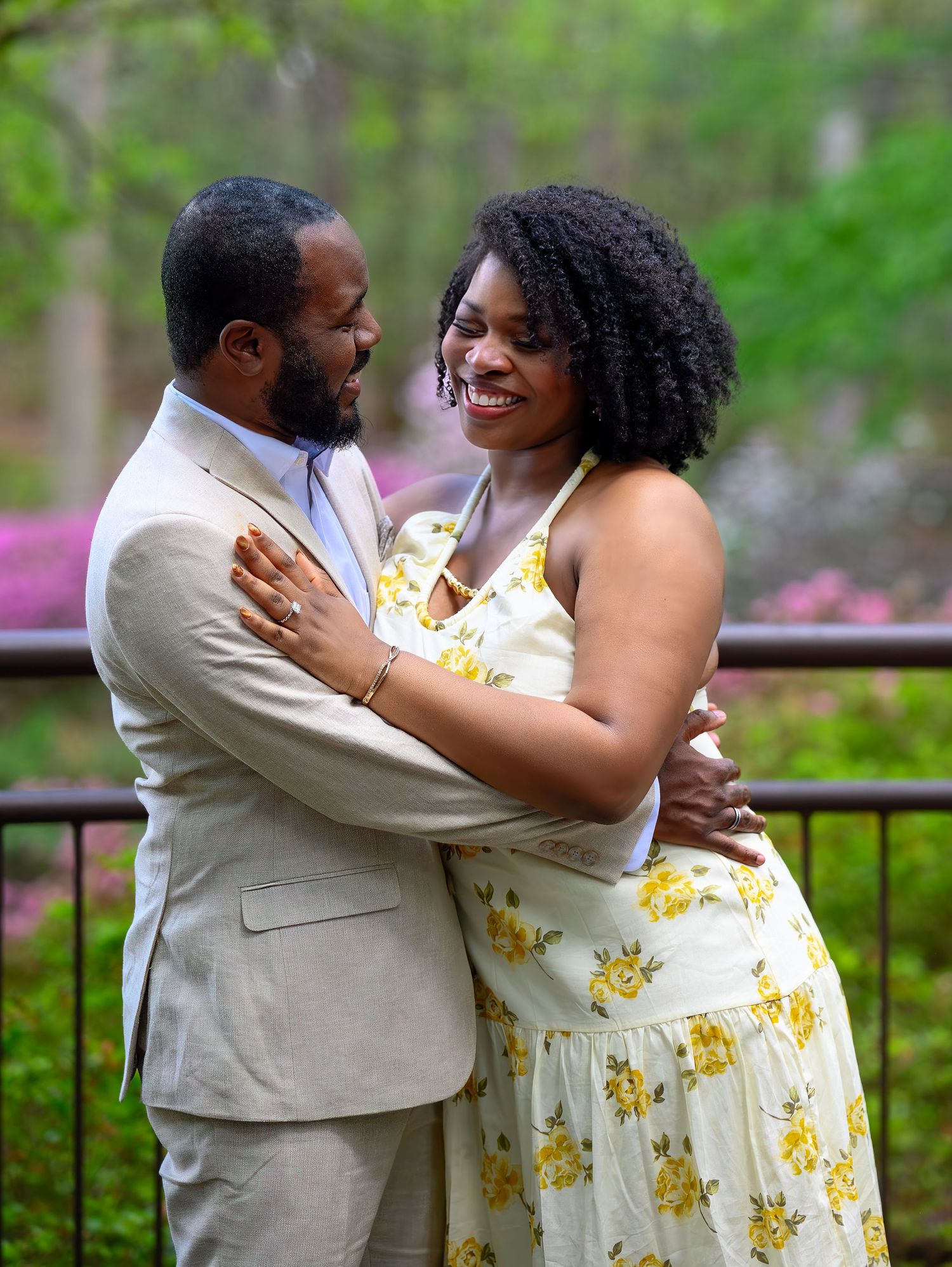 Happy engaged couple embracing and smiling at each other on a garden terrace with pink flowers in background.