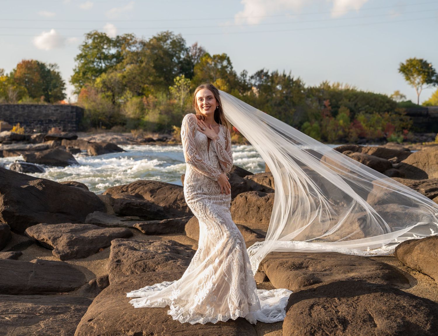 A white lace gown and veil flow gracefully in the wind on a rocky shoreline during golden hour.