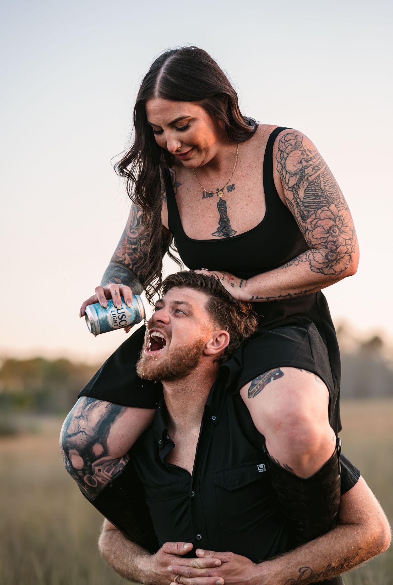 Playful couple shares joyful laughter while embracing in an outdoor field at sunset.