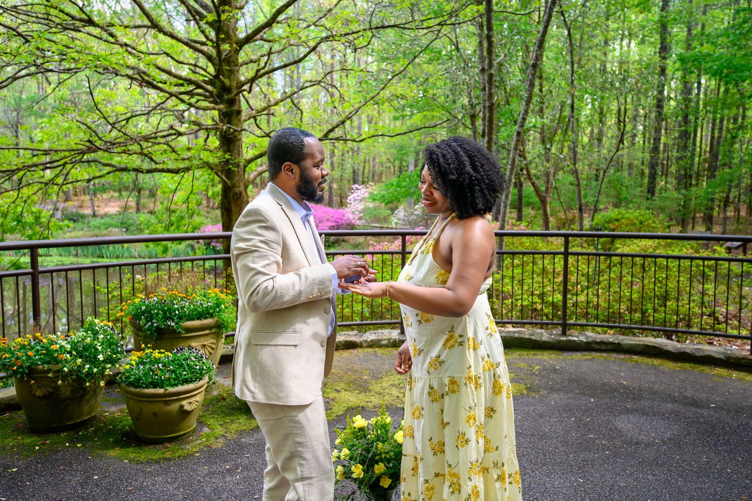 A couple shares a romantic moment in a lush green garden with blooming azaleas, exchanging rings during a proposal.