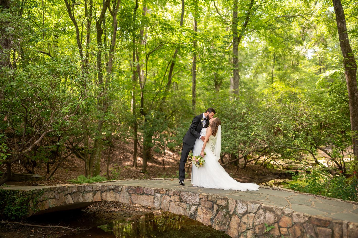 A couple stands together on a stone bridge in a lush green forest setting during a wedding ceremony.