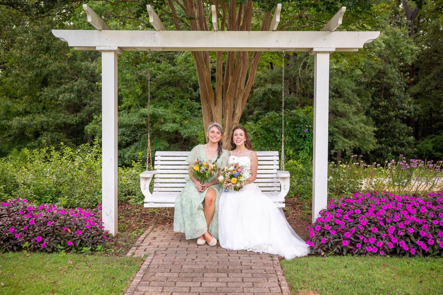 Two people sit on a white swing under a garden pergola surrounded by vibrant purple flowers and greenery.