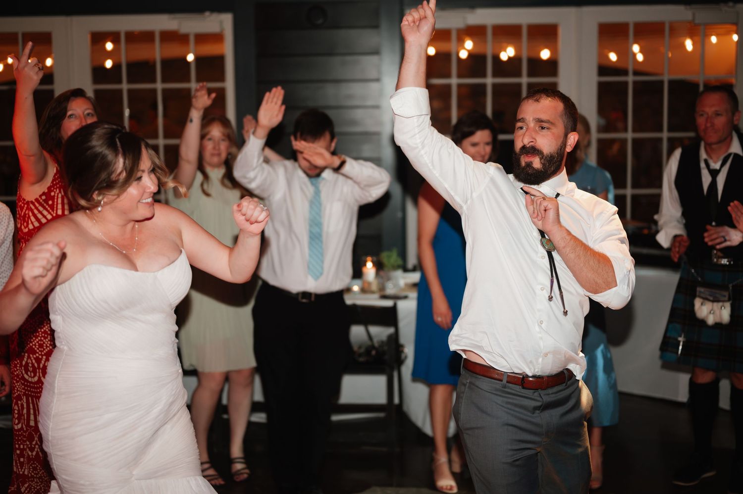 Wedding guests dance enthusiastically at a reception with warm string lights in the background.