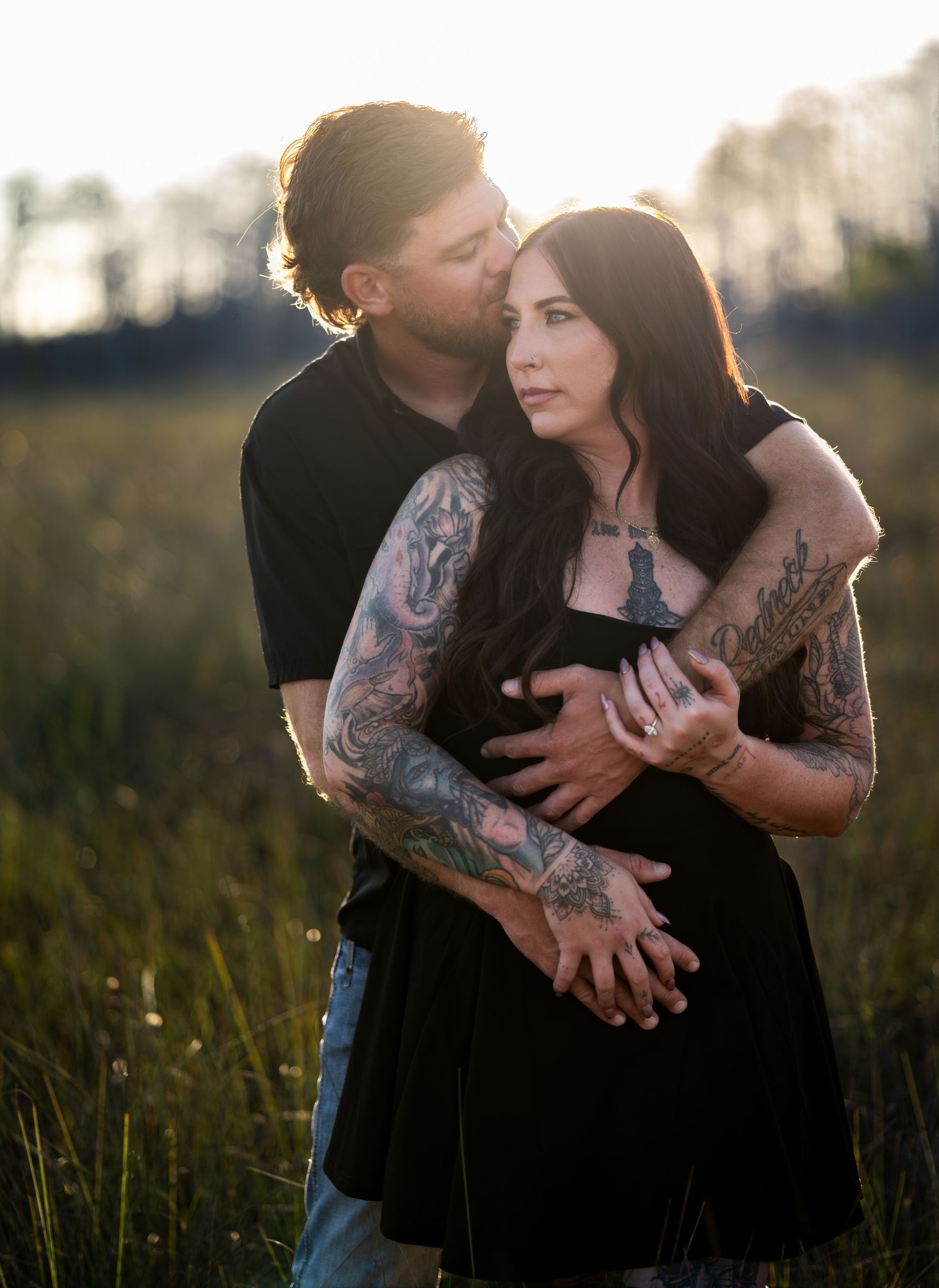 Couple embraces tenderly in tall grass with sun flare and natural lighting.