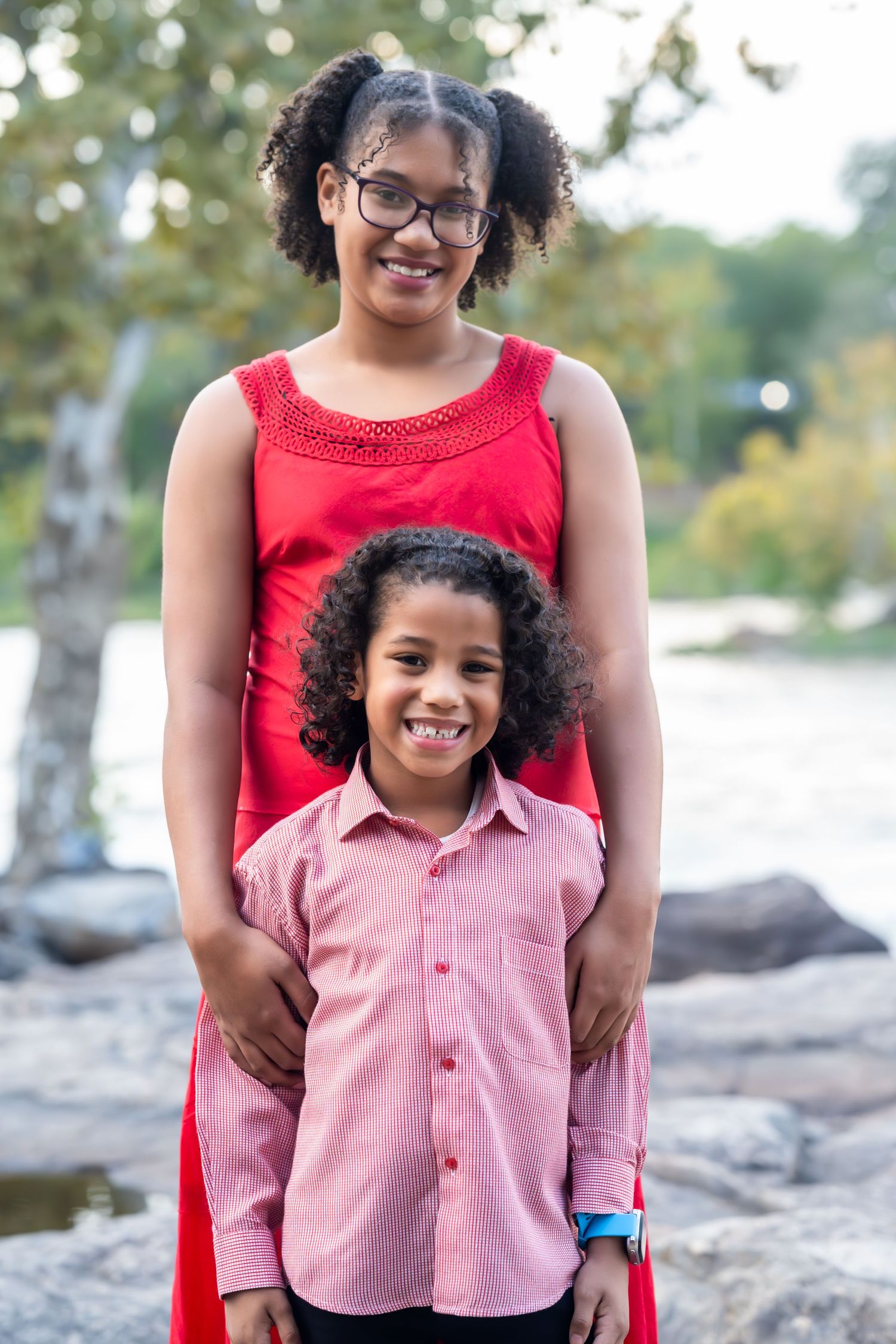 A parent and child in red and pink outfits share a sweet moment on rocky terrain by the water.