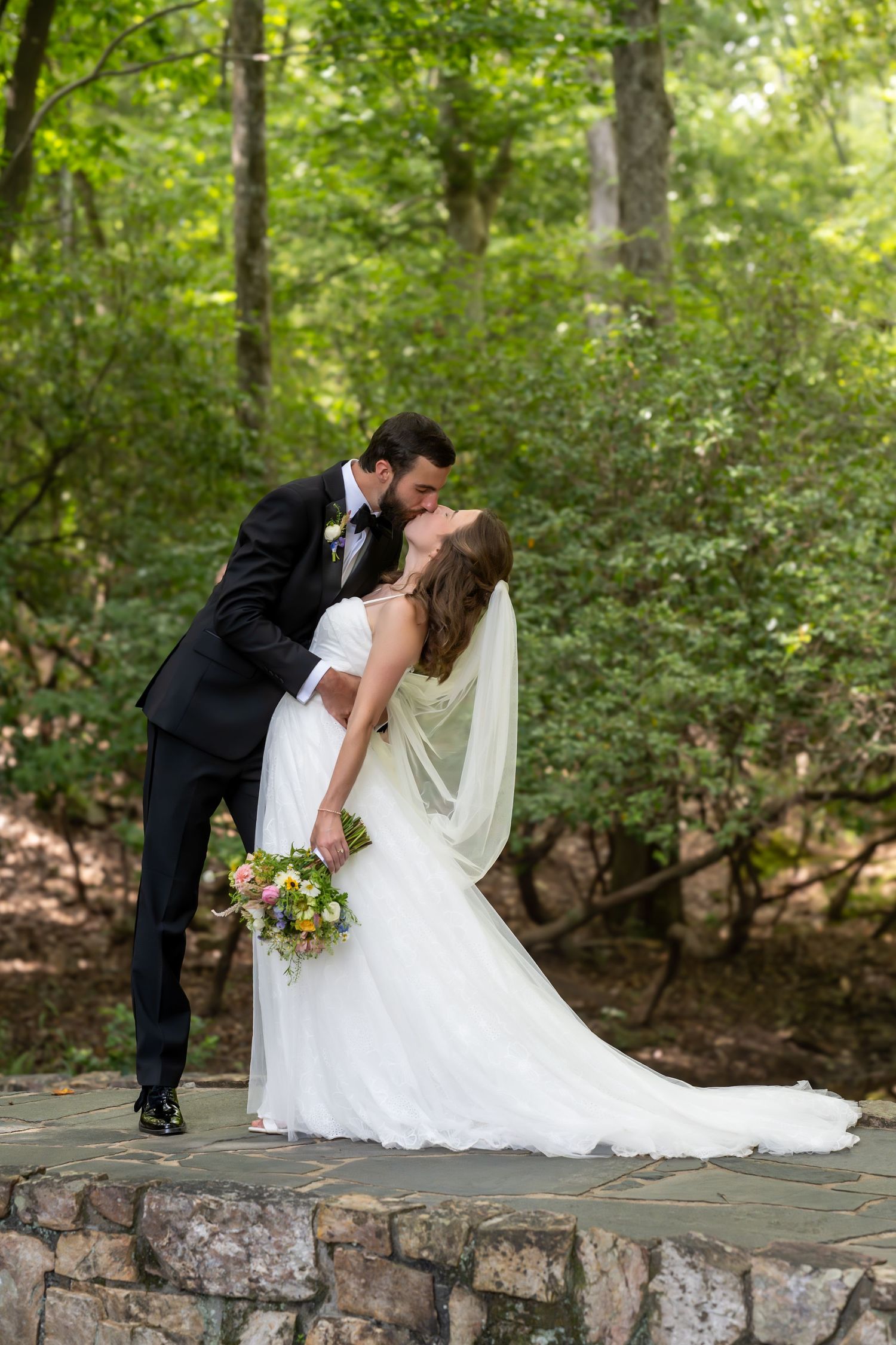 A romantic dip kiss between newlyweds on a stone wall surrounded by lush green forest scenery.