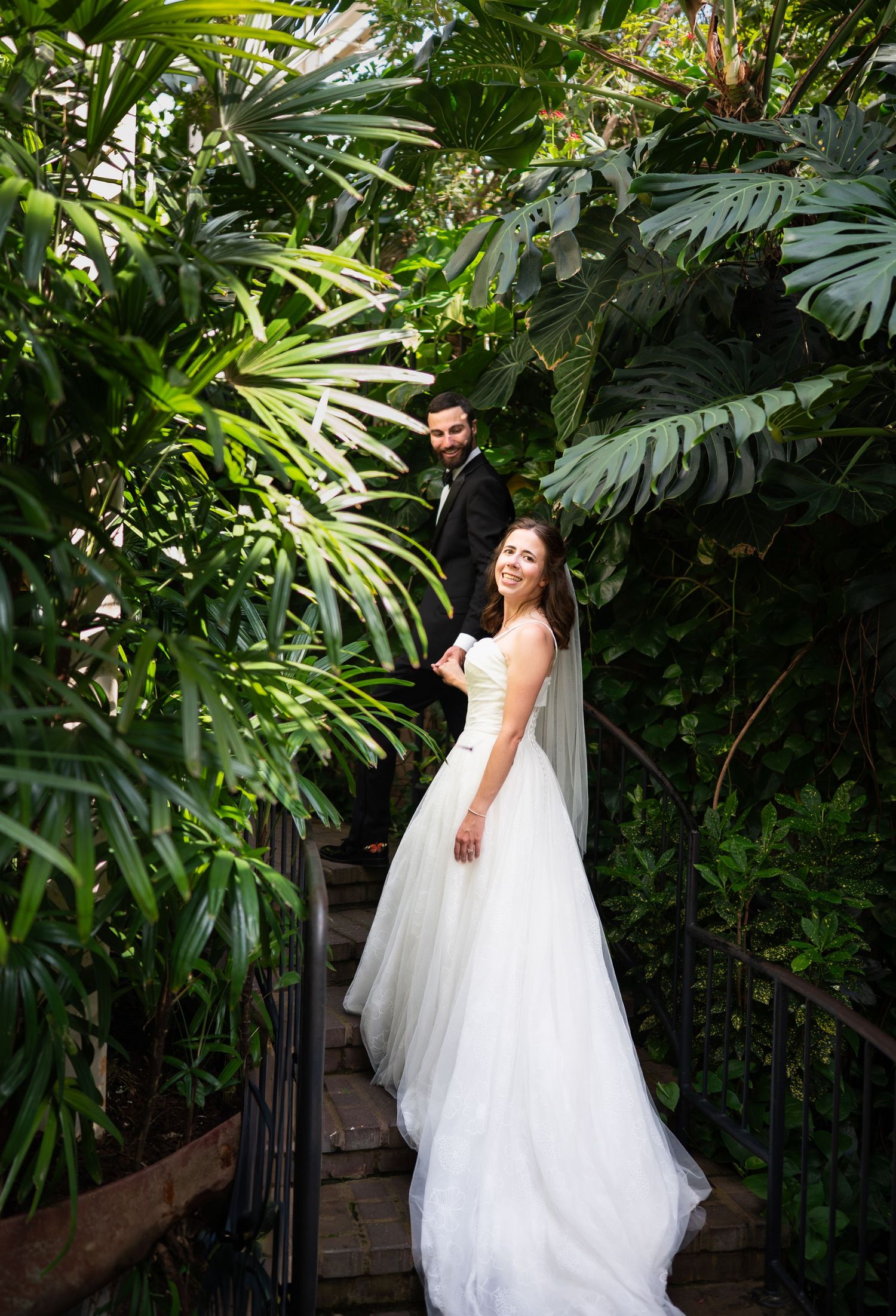 A bride in a flowing white wedding gown poses elegantly among lush tropical foliage in a garden setting.