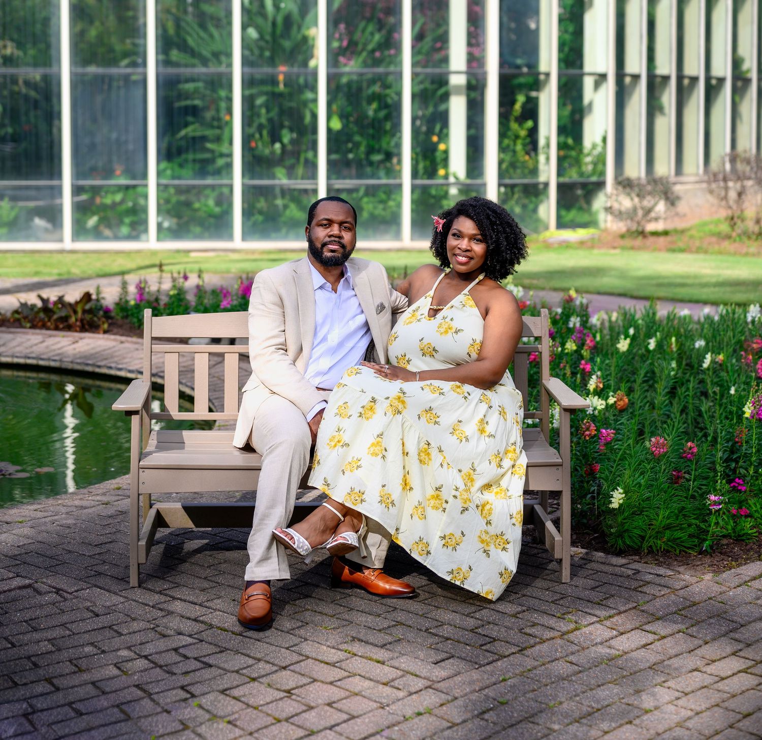 Couple posing on a brick garden bench near a greenhouse, woman in yellow floral dress, man in beige suit.