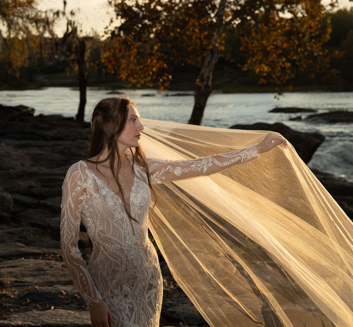 Golden sunlight illuminates bride's flowing veil as she poses near rocky shoreline at sunset.