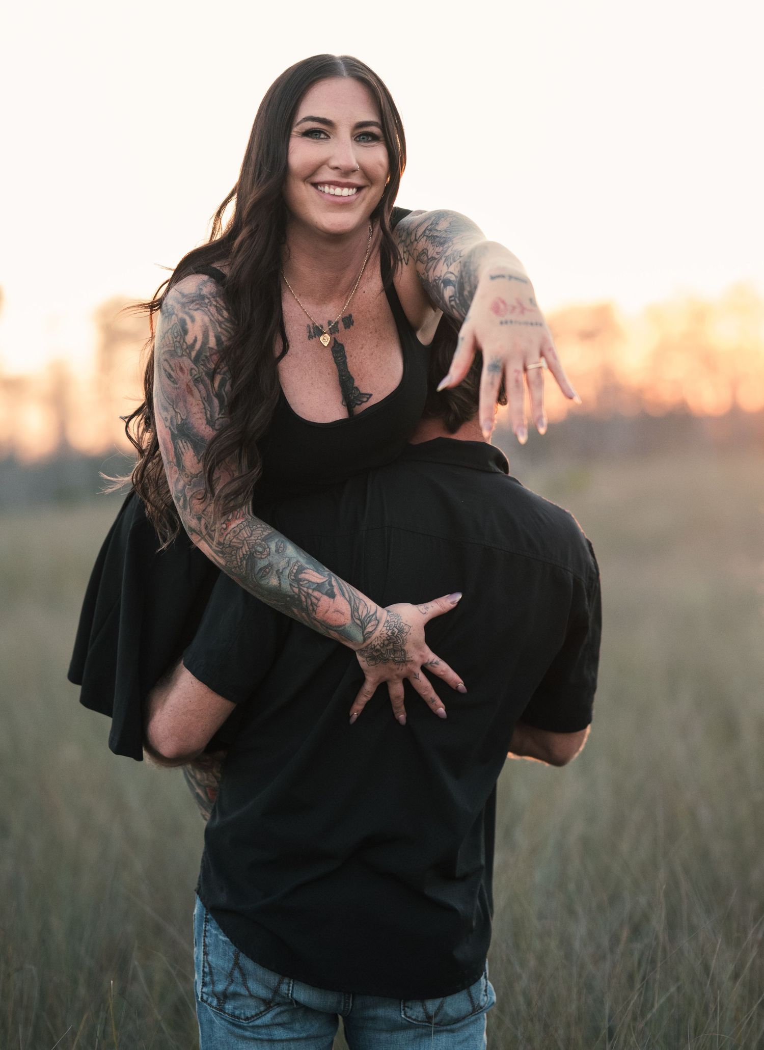 A couple shares a playful moment in a field as the sun sets, with one person being lifted up in laughter.