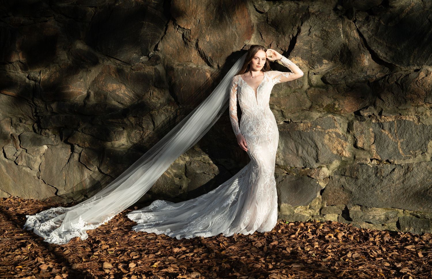Dramatic bridal portrait with flowing veil against textured stone wall and fallen autumn leaves.