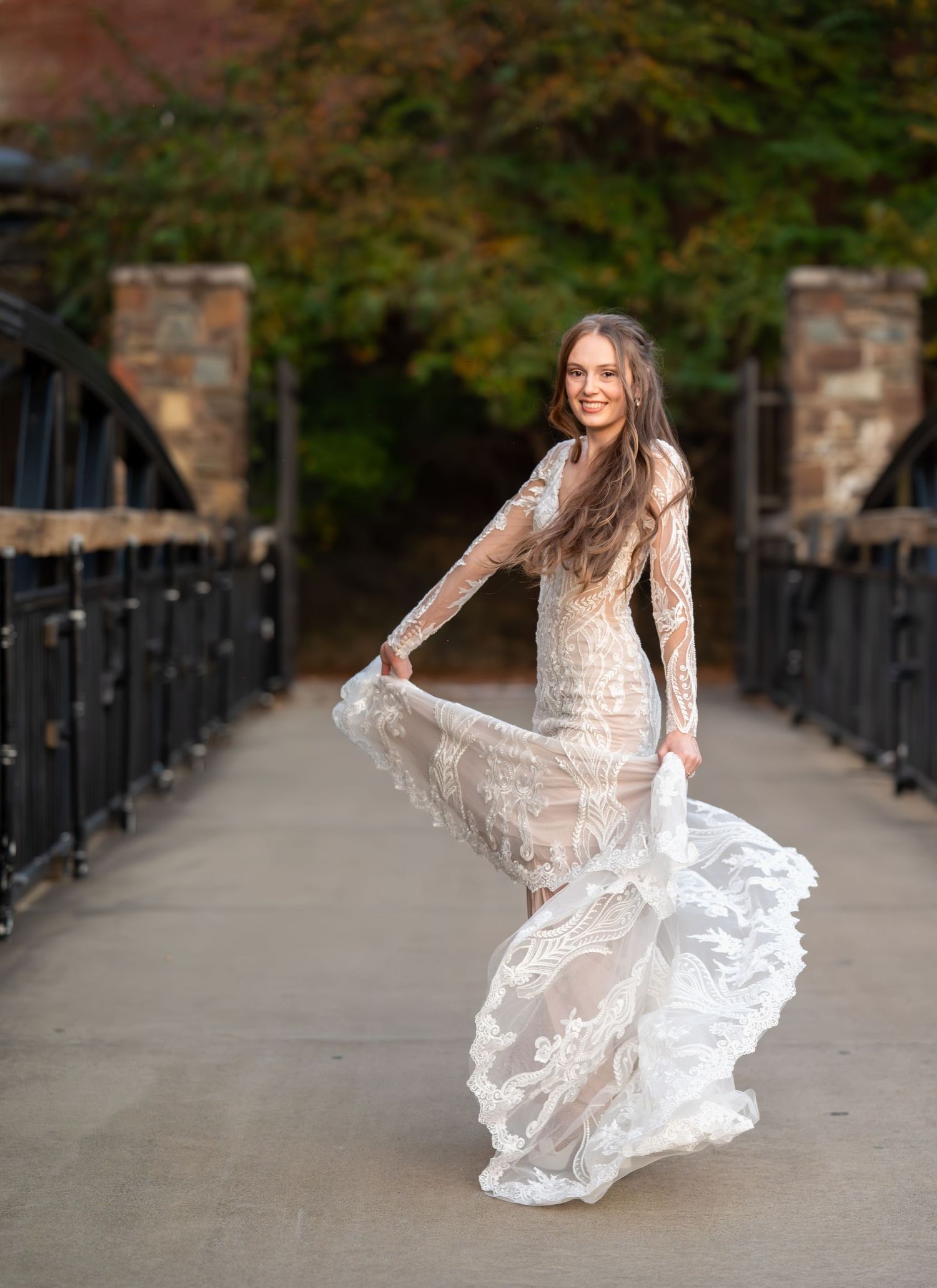 An elegant bride twirls in a sheer lace wedding gown along a tree-lined pathway.