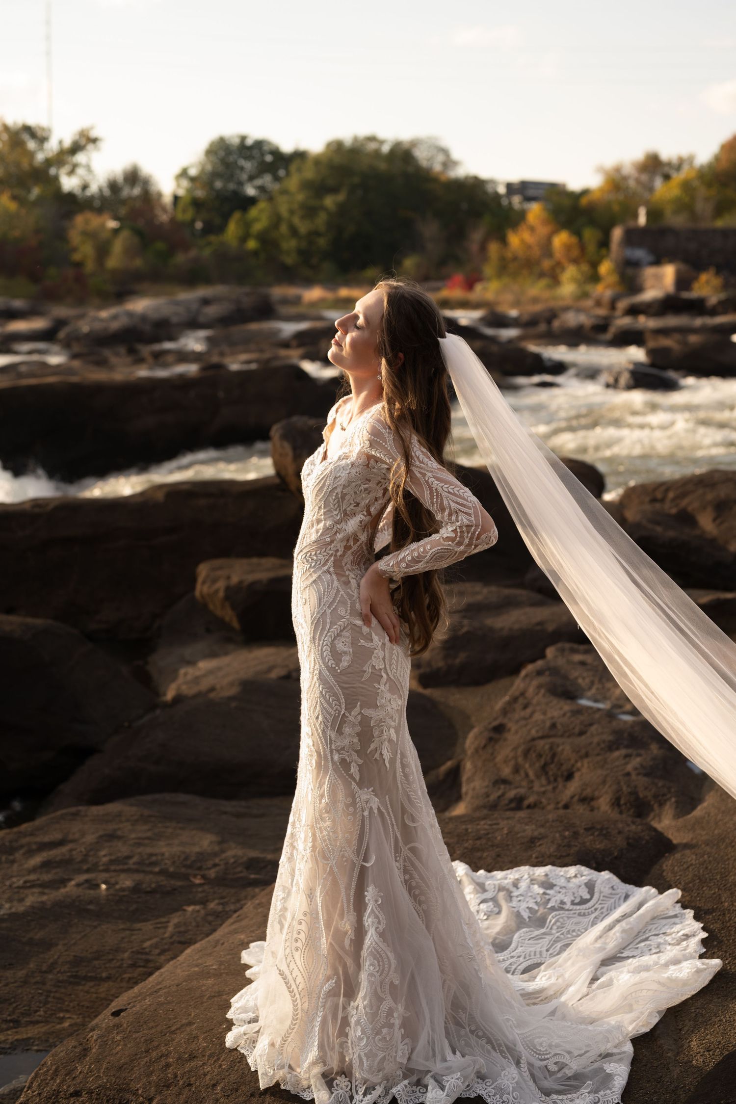 Elegant lace wedding dress with flowing train photographed against rocky riverside landscape at sunset.